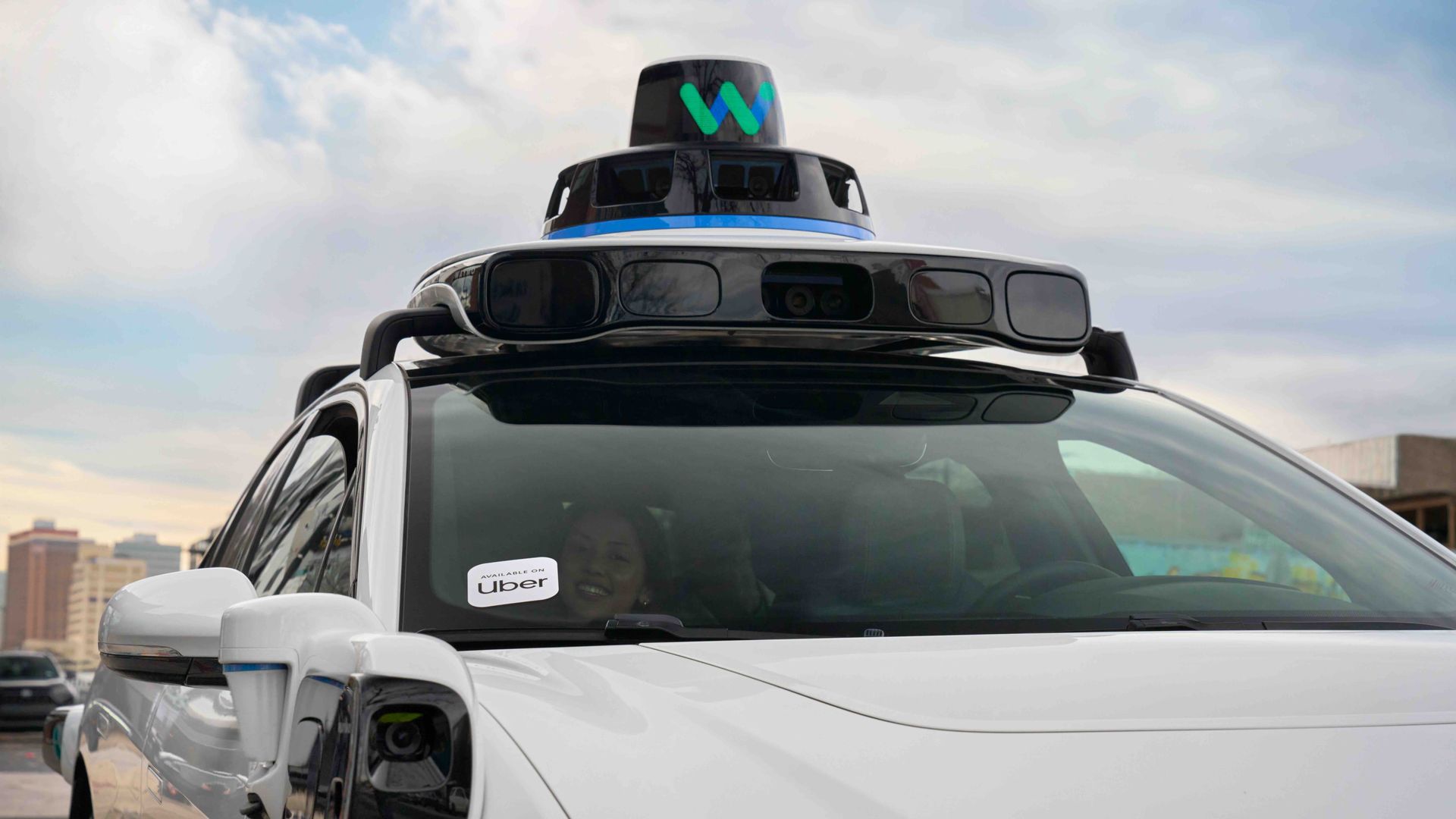A photo of a person in the front of a Waymo. The car has a sticker on the windshield that reads "Uber."