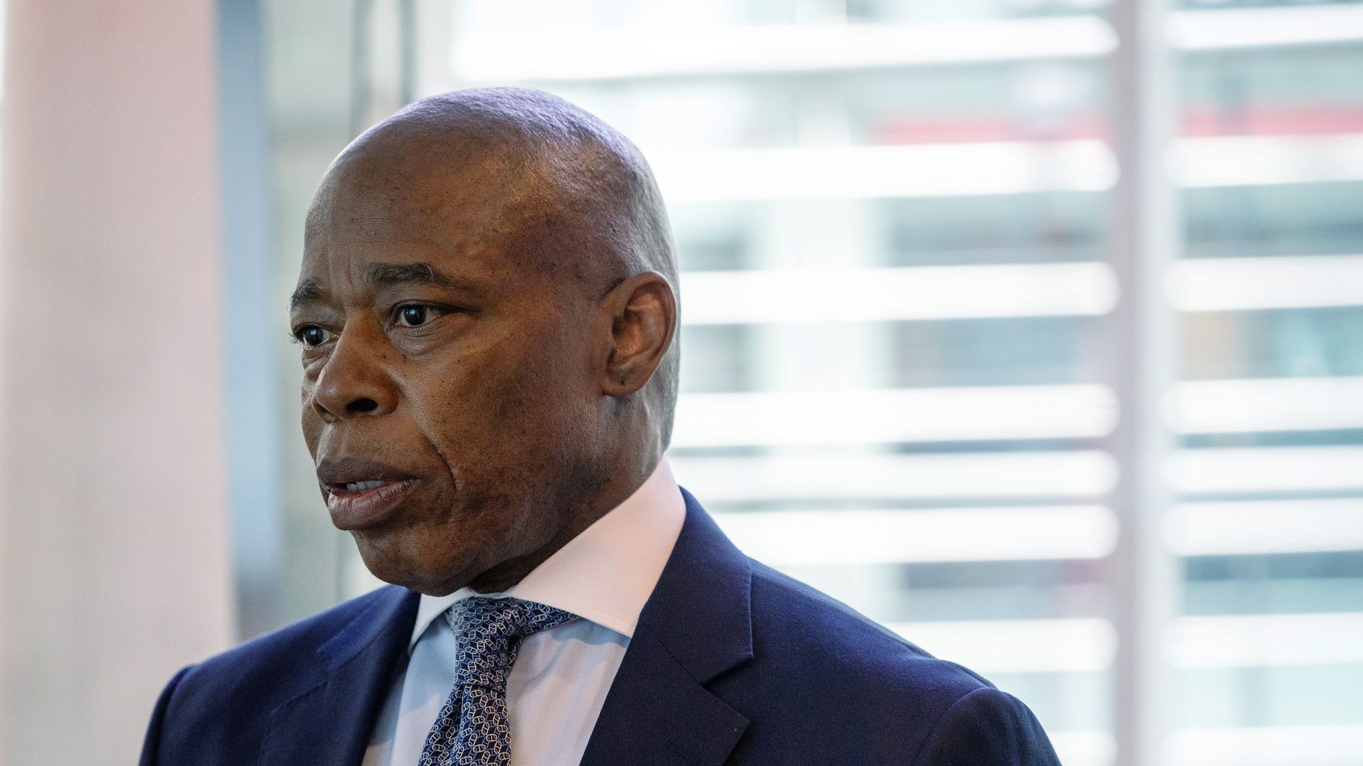 Portrait of a man with a shaved head wearing a navy blue suit, white shirt, and patterned blue tie, with a window featuring horizontal blinds in the background.