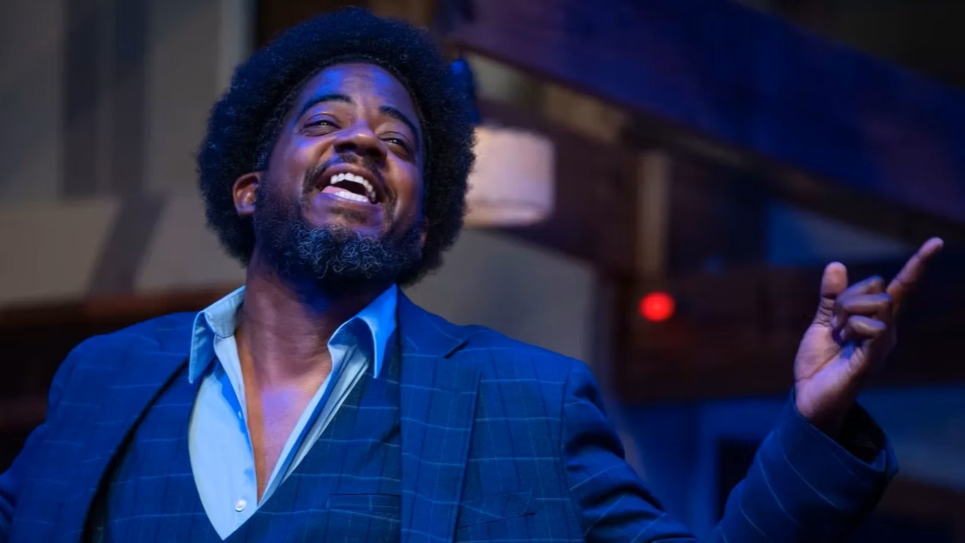 A joyful man with an afro and beard, wearing a blue pinstripe suit and light blue shirt, raises his right hand and smiles on stage under blue lighting.