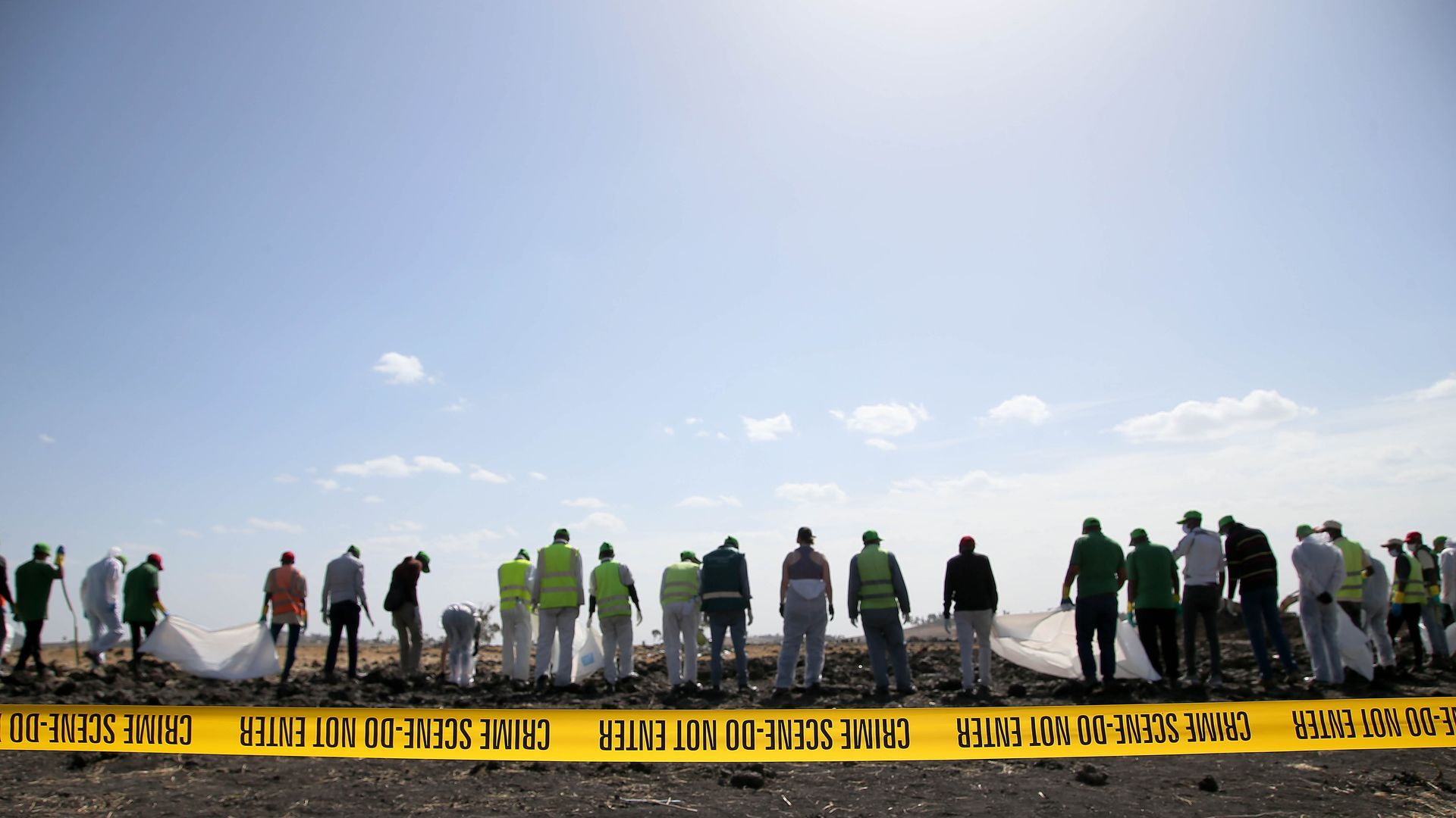A line of investigators in yellow vests clean up a plane wreckage site. 