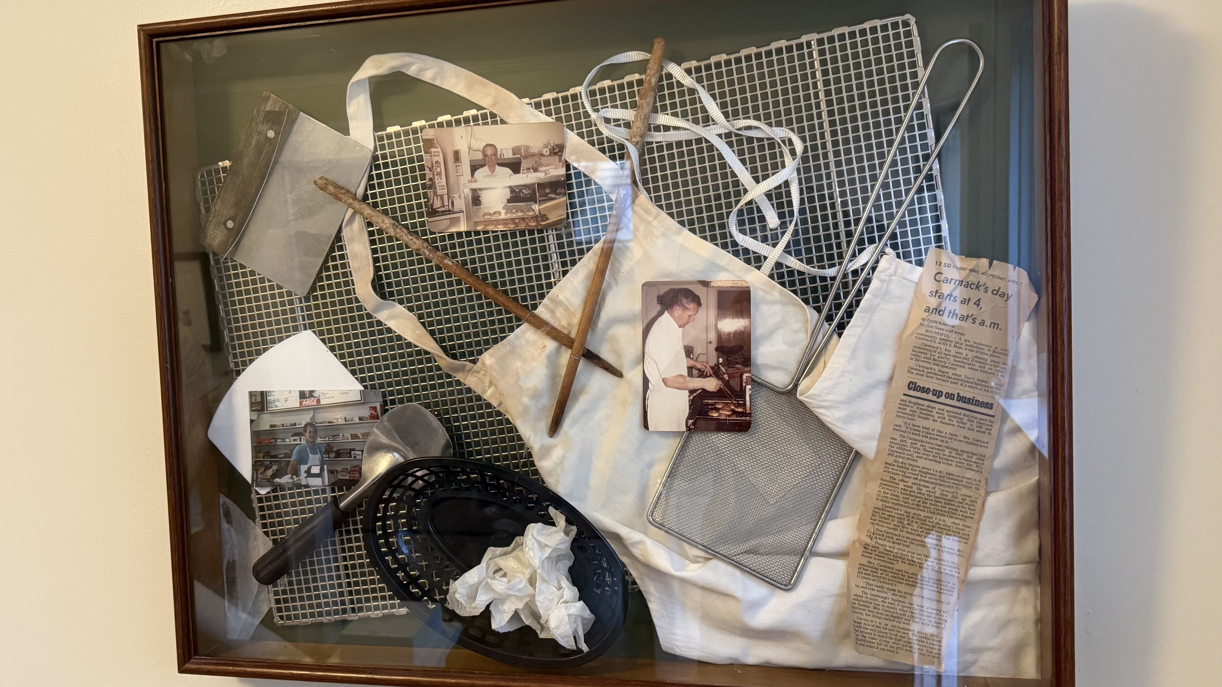 Framed display with a white apron on a metal grid, vintage cooking photos, a wooden stick, tongs, a wire rack, a newspaper clipping, and a black basket with crumpled paper.