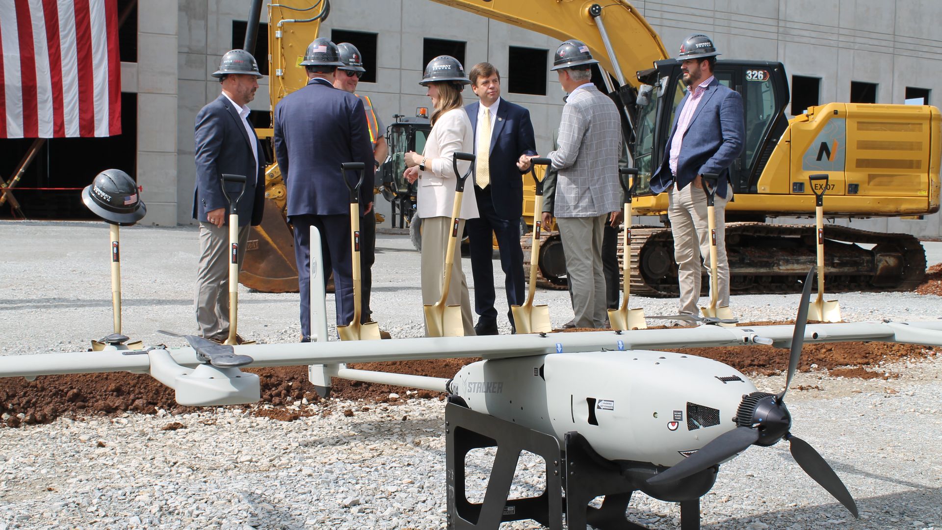 Group of suited professionals wearing hard hats stand with shovels at a construction site, a large yellow excavator behind them, and an American flag visible on the left.