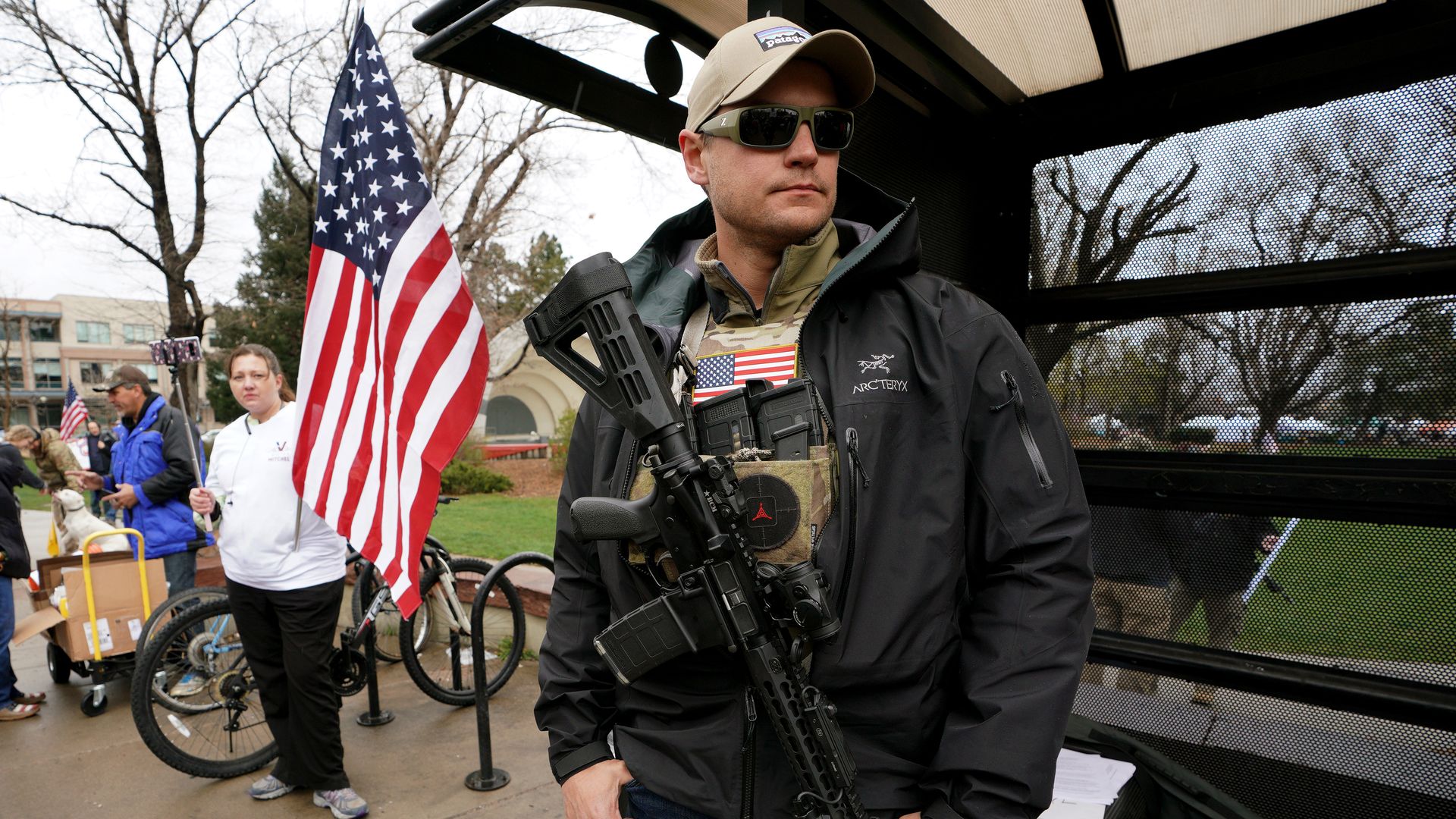 Man stands with an AR-15 rifle slung across his chest with an American flag in the background