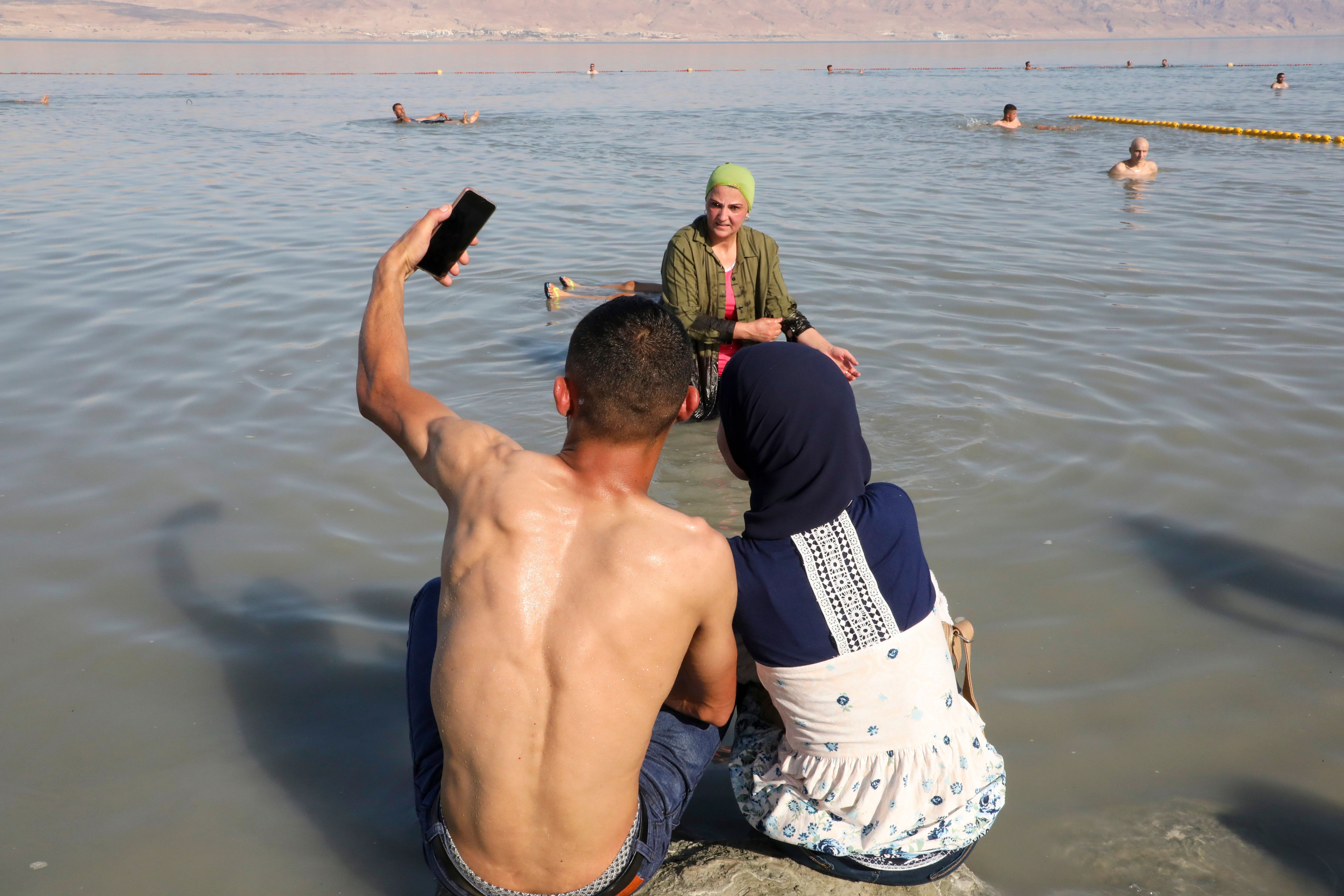 A shirtless man and woman take a selfie while sitting on a beach