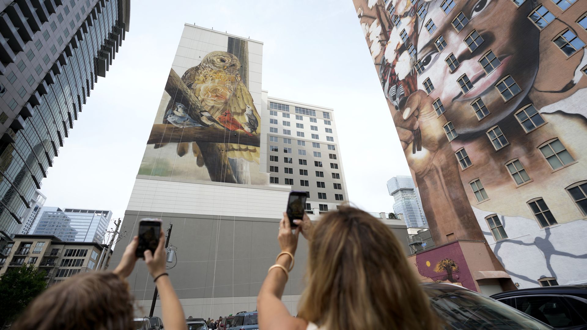 Low-angle city scene with tall buildings displaying murals: a giant golden bird on a white tower and a large woman's face on the adjacent wall, while people in the foreground snap photos.
