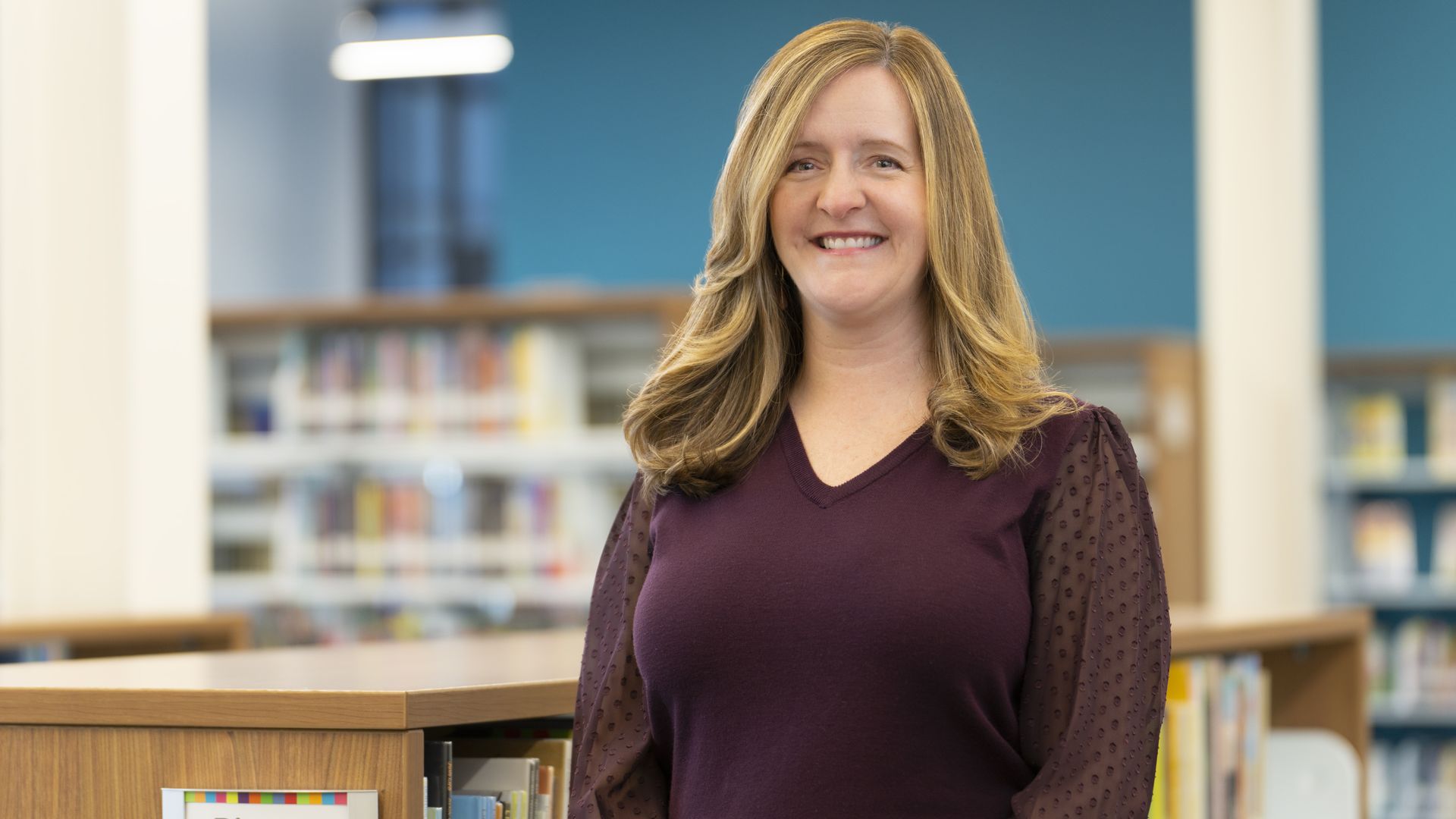 Smiling woman with shoulder-length blonde hair in a maroon top stands in a library; blue walls and blurred bookshelves behind her, with a wooden shelf in the foreground labeled Picture Books.