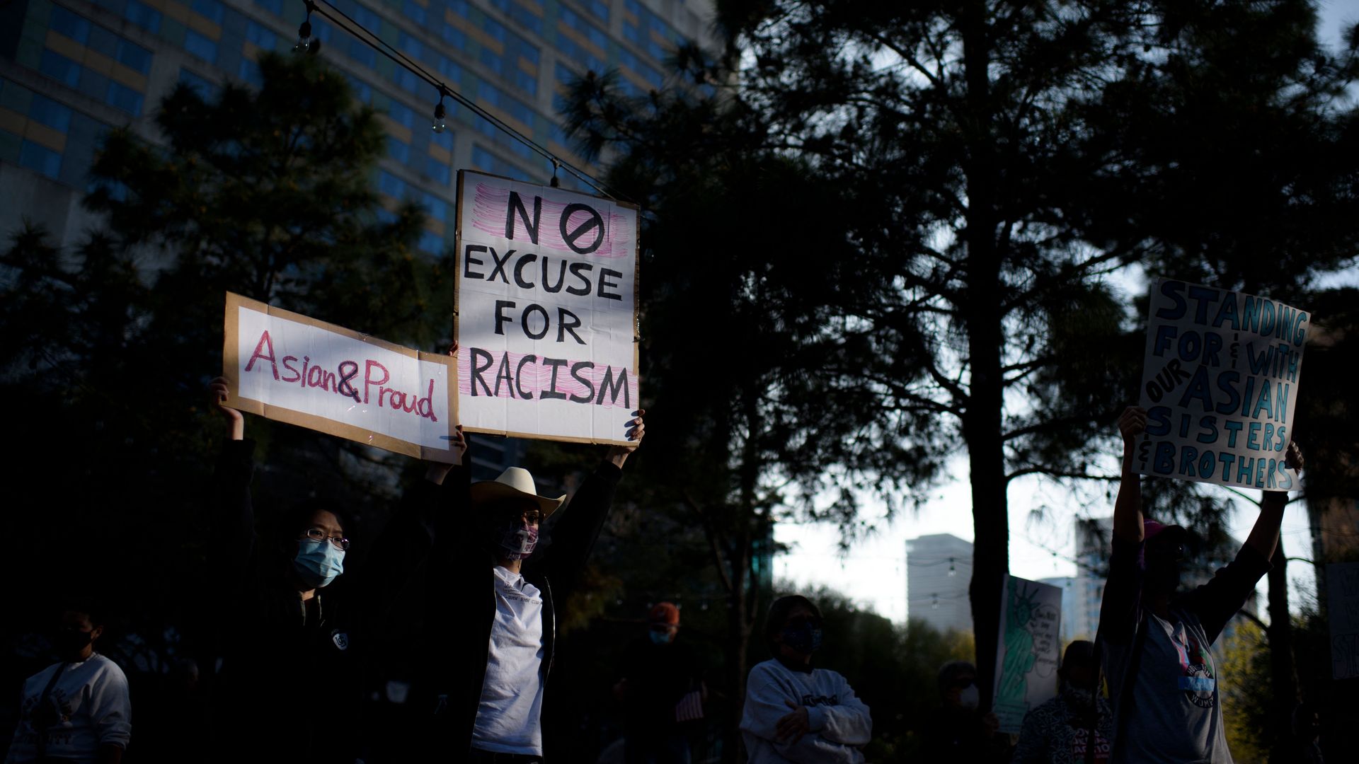 Photo of two protesters, one holds a sign that says "Asian&Proud" and the other holds a sign that says "No excuse for racism"