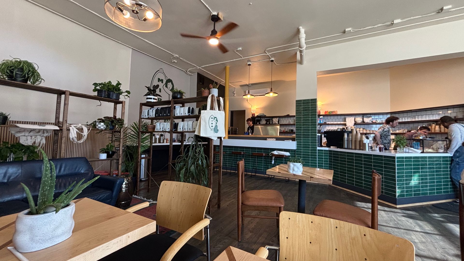 Bright cafe interior with wooden tables and chairs, green tiled counter, shelves of plants and mugs, exposed white pipes on a gray ceiling, a ceiling fan and hanging lights, customers in the back.