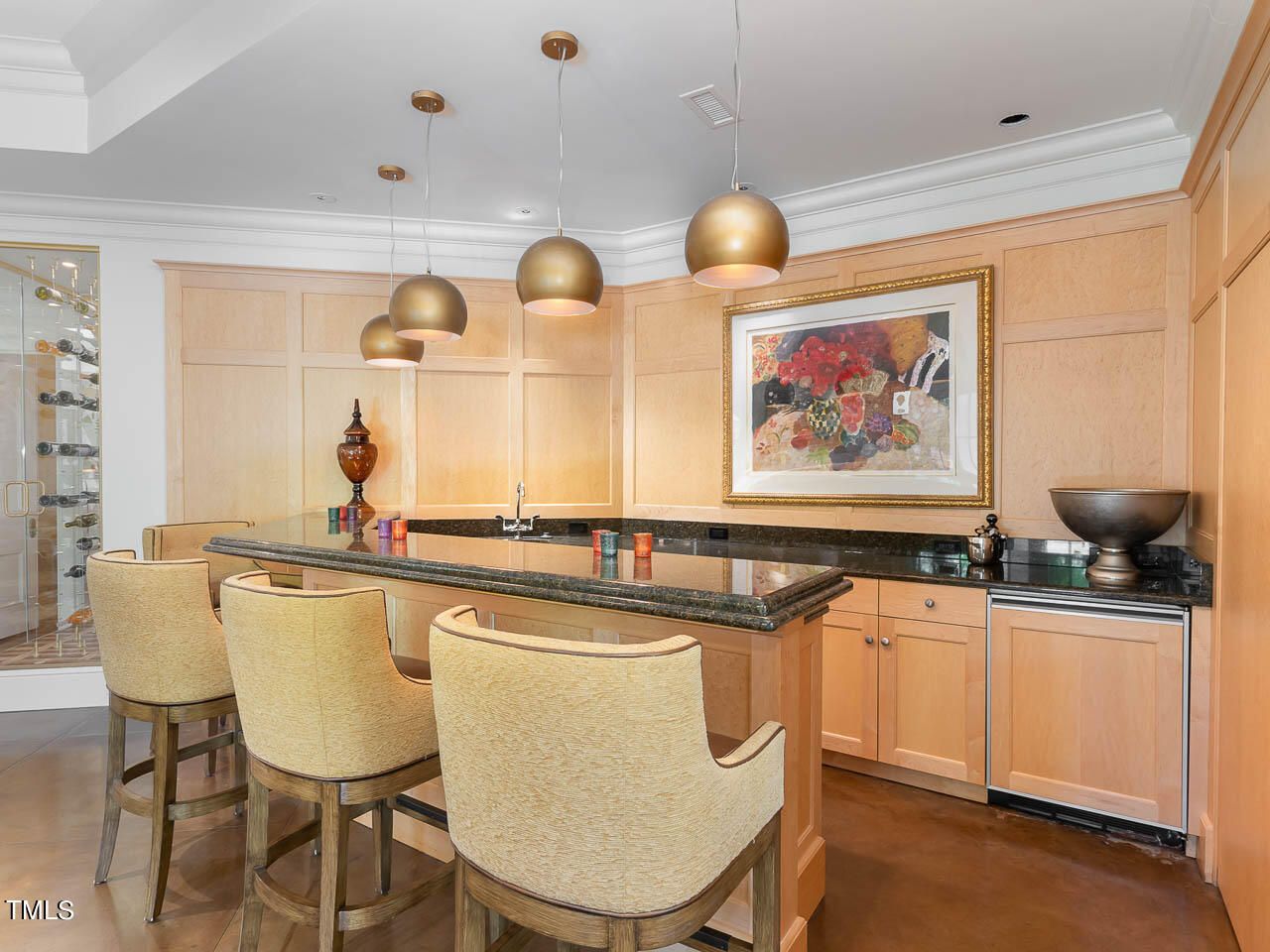 Modern home bar with beige cushioned stools, wooden cabinetry, black granite countertops, gold pendant lights, wine rack, and framed floral artwork on light wood paneled walls.