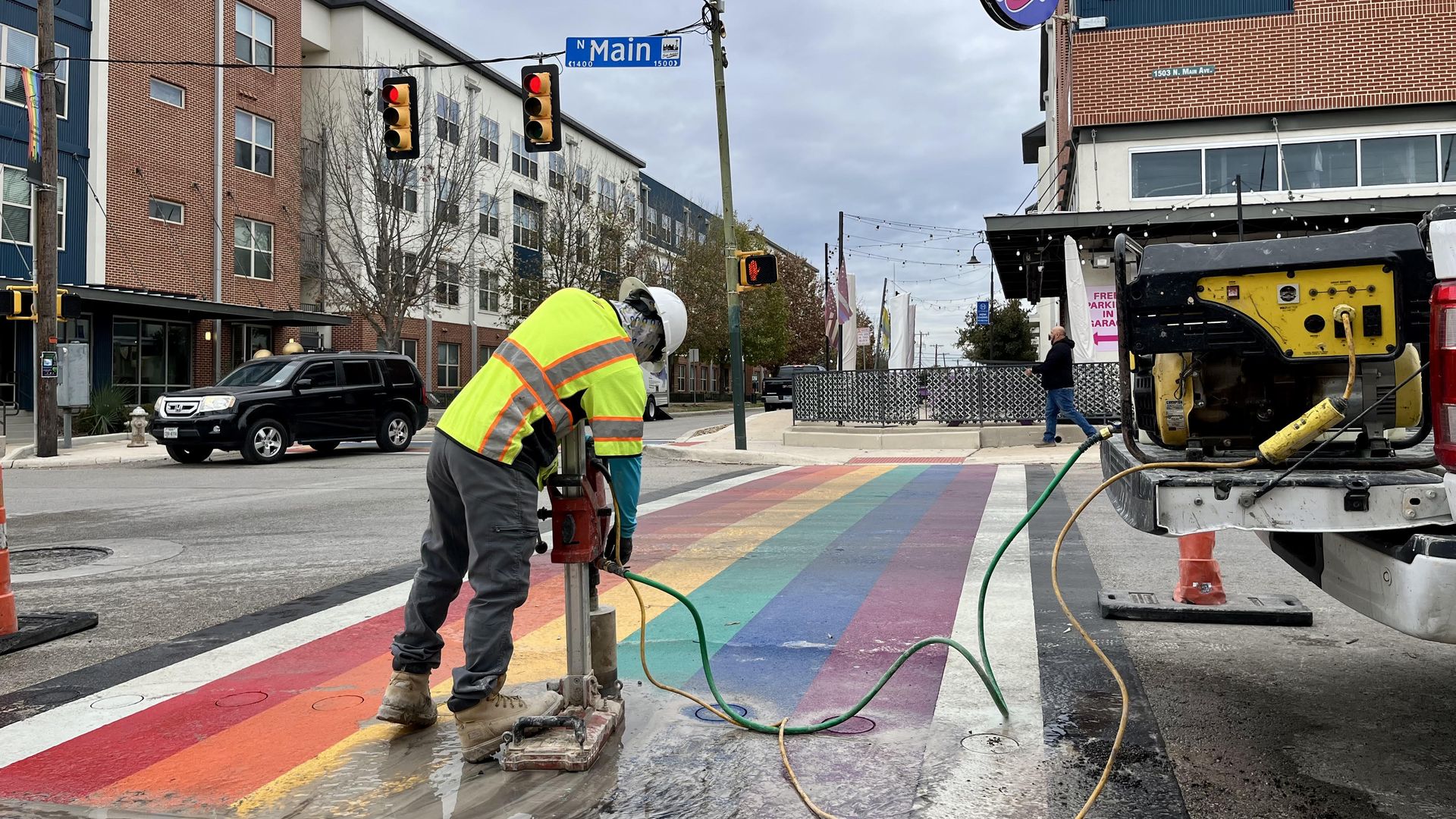 A construction worker in a yellow reflective jacket and helmet operating equipment on the rainbow-colored crosswalk at North Main Avenue and Evergreen Street. Red traffic lights and brick buildings stand in the background.