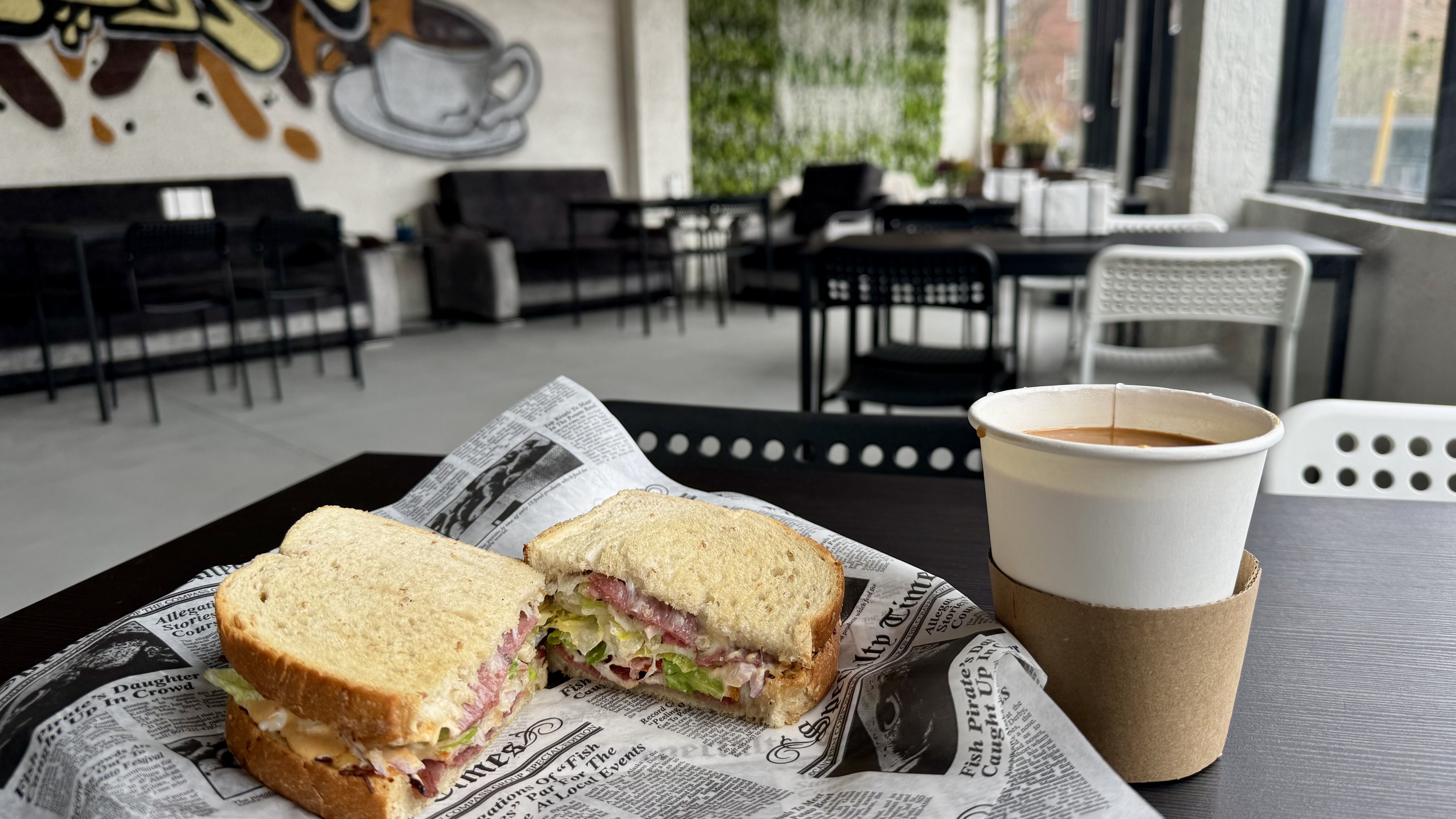 Sandwich on newspaper with a coffee cup on a table, in an outdoor cafe with black and white chairs and graffiti of a coffee mug on the wall behind.