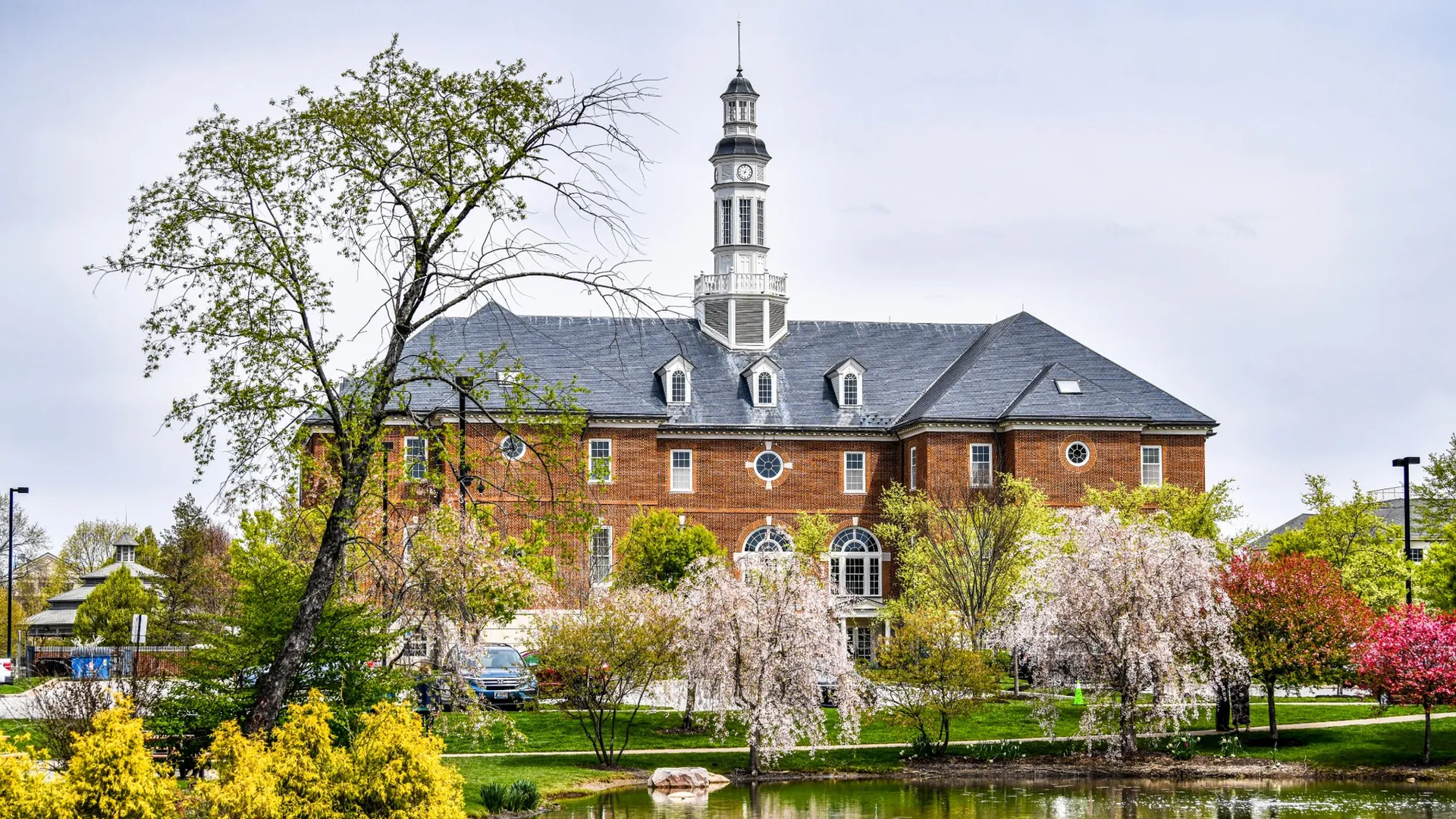 A red brick building with a tall white clock tower and slate roof, flanked by blooming trees and shrubs; a small pond in the foreground reflects the scene.