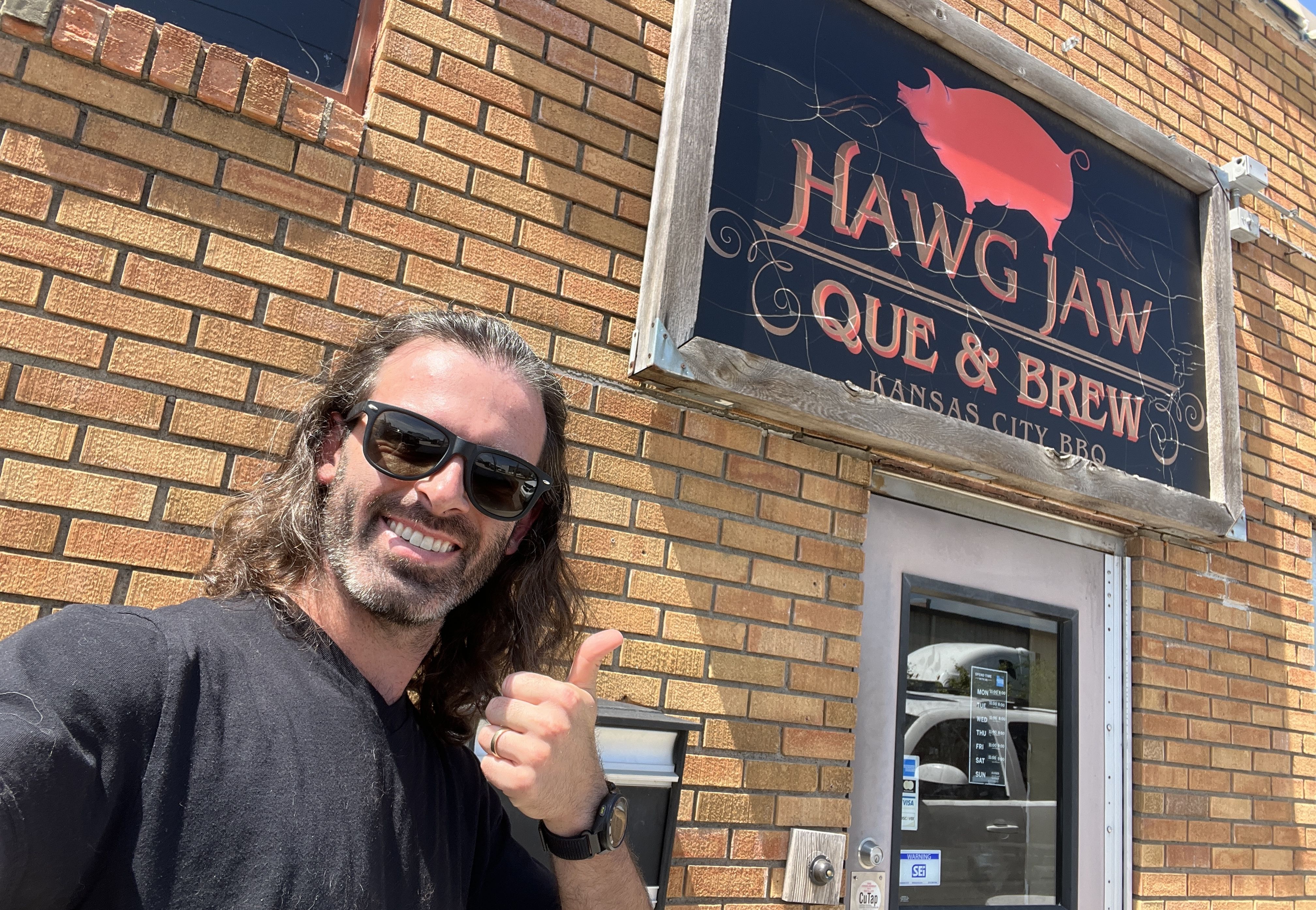 Reporter Travis Meier giving thumbs up in front of brick building with a sign that reads "HAWG JAW QUE & BREW Kansas City BBQ" featuring a red pig silhouette.