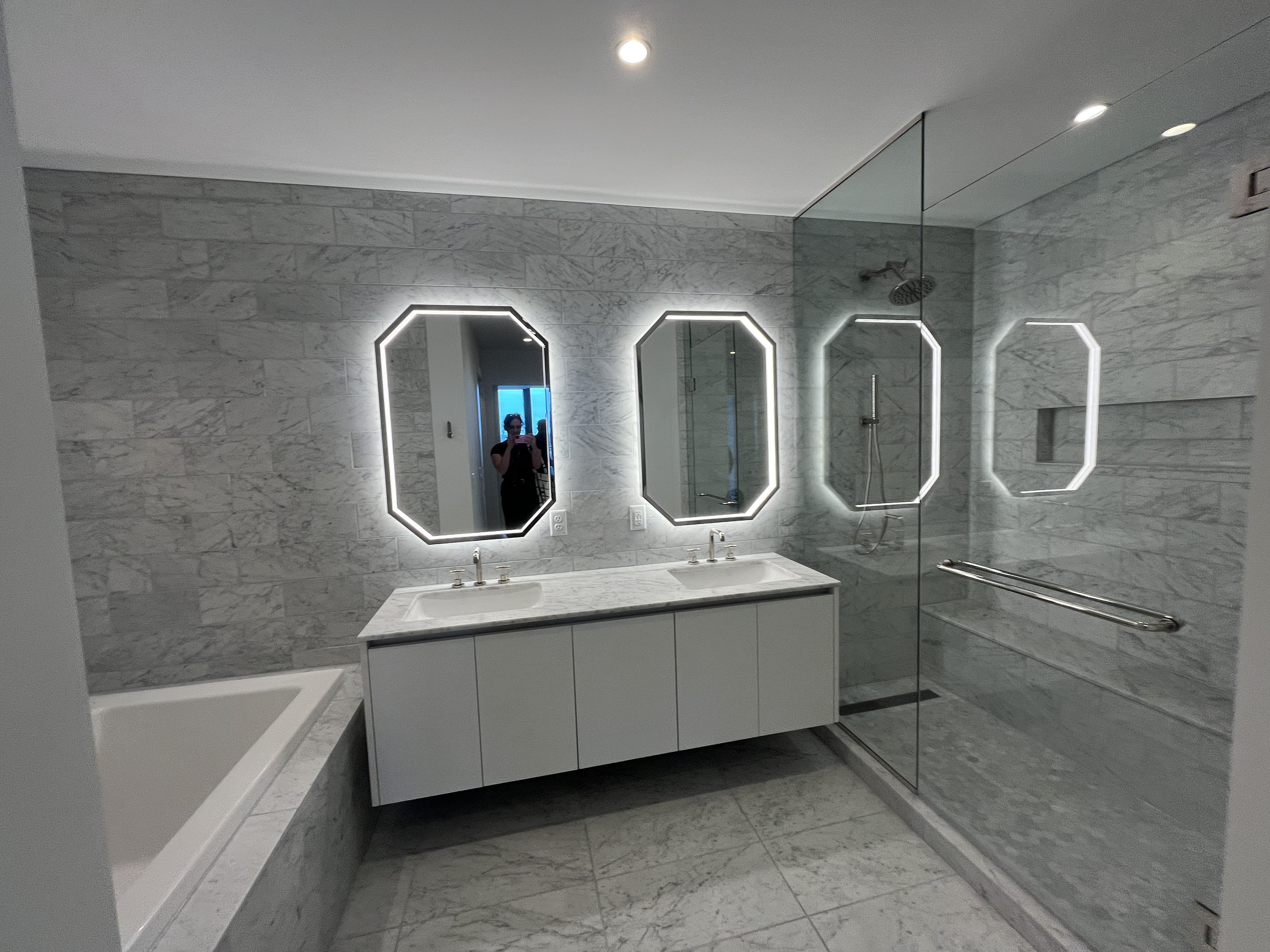 Modern marble bathroom with a double vanity, backlit hexagonal mirrors, a bathtub on the left, and a glass shower enclosure with a rainfall head; a person is visible taking a photo in the mirror.