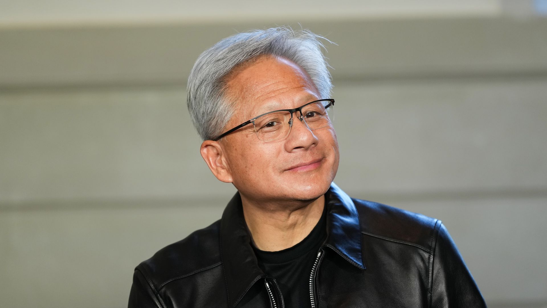 Portrait of a man with silver hair and rectangular glasses, wearing a black leather jacket, looking to the right with a slight smile against a neutral pale background.