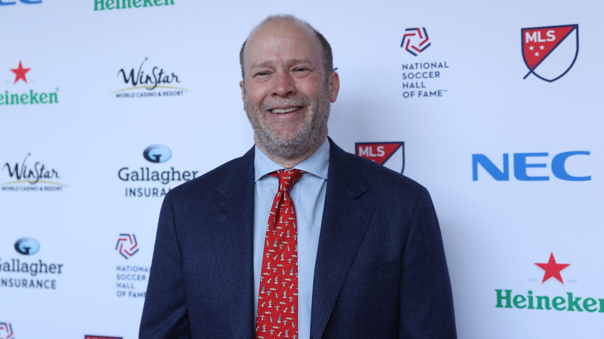 A man wearing a suit stands in front of a white step and repeat