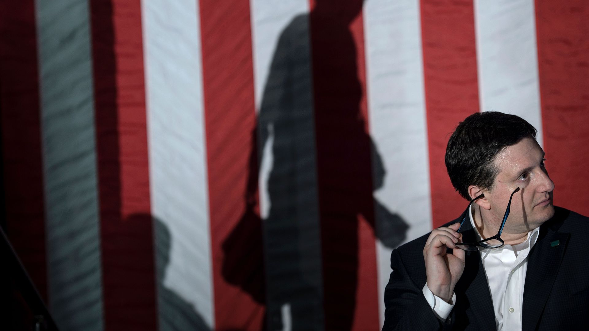 Philippe Reines sits and holds his glasses to his face with a large American flag behind him, and the shadow of Hillary Clinton walking on stage.