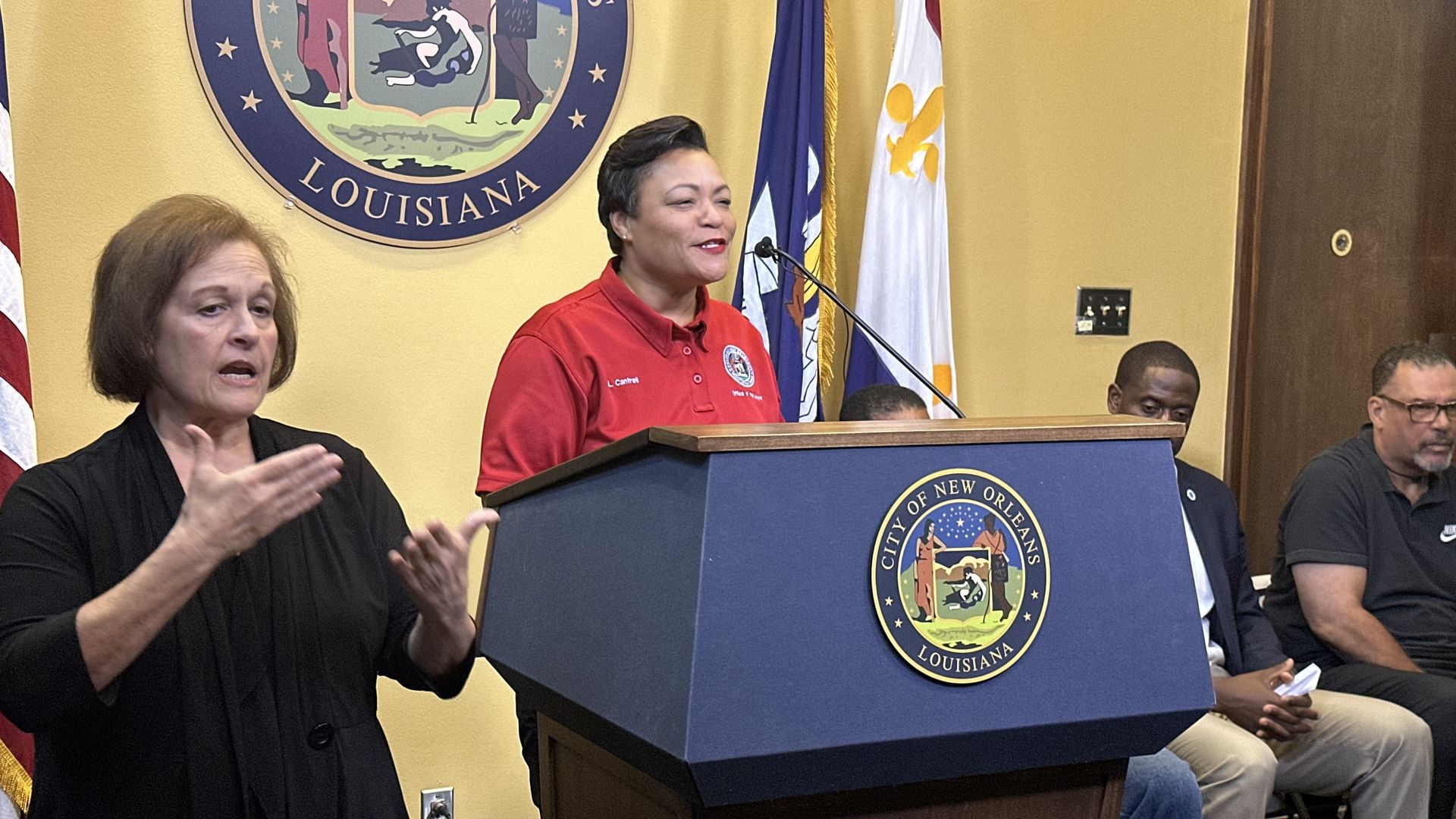 Mayor LaToya Cantrell stands at a podium and speaks. A sign language interpreter stands next to her.