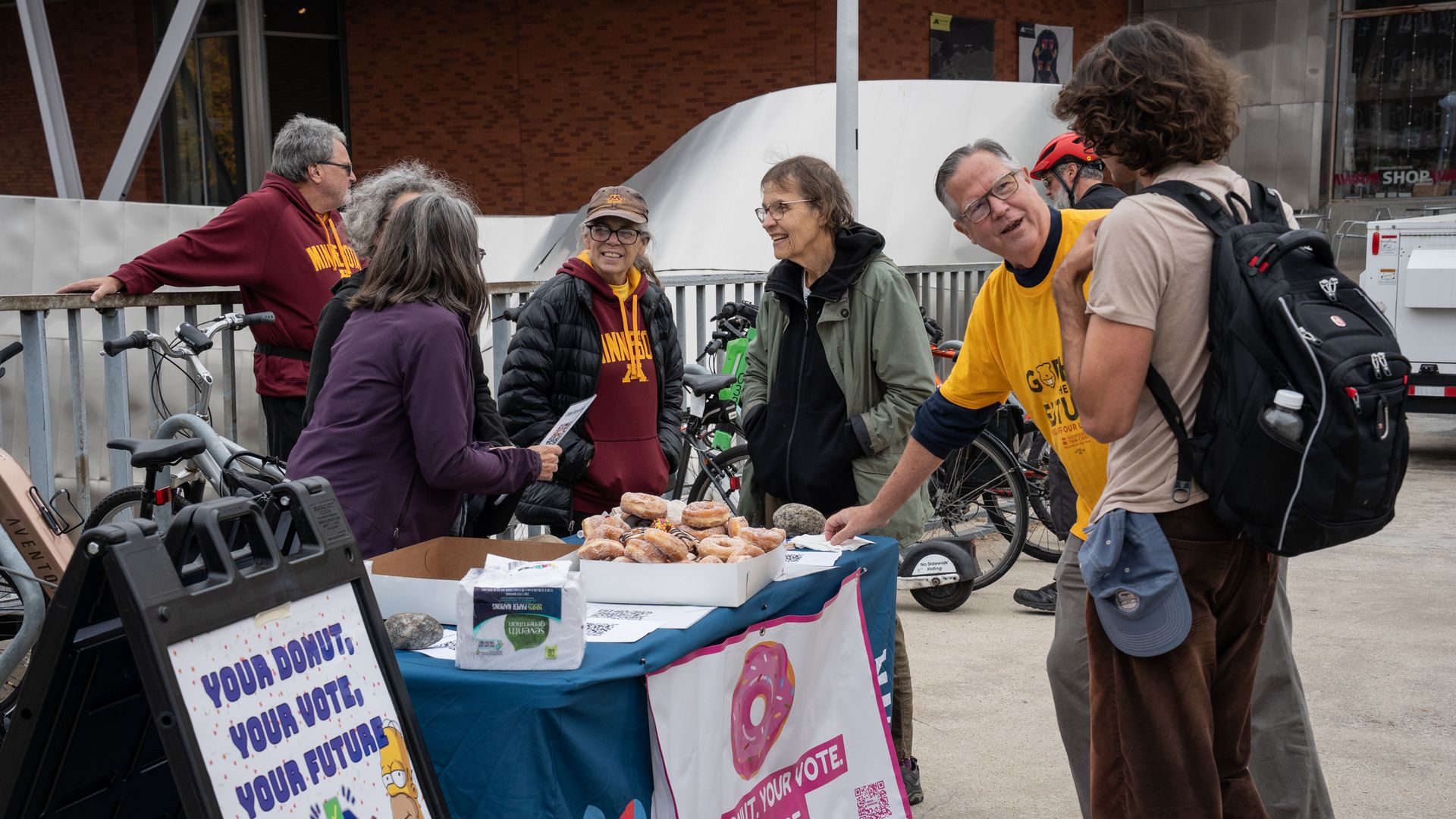 Group of adults gathers around a table with donuts and vote posters reading Your Vote Your Future; cyclists and bikes lean against a railing in an urban setting.