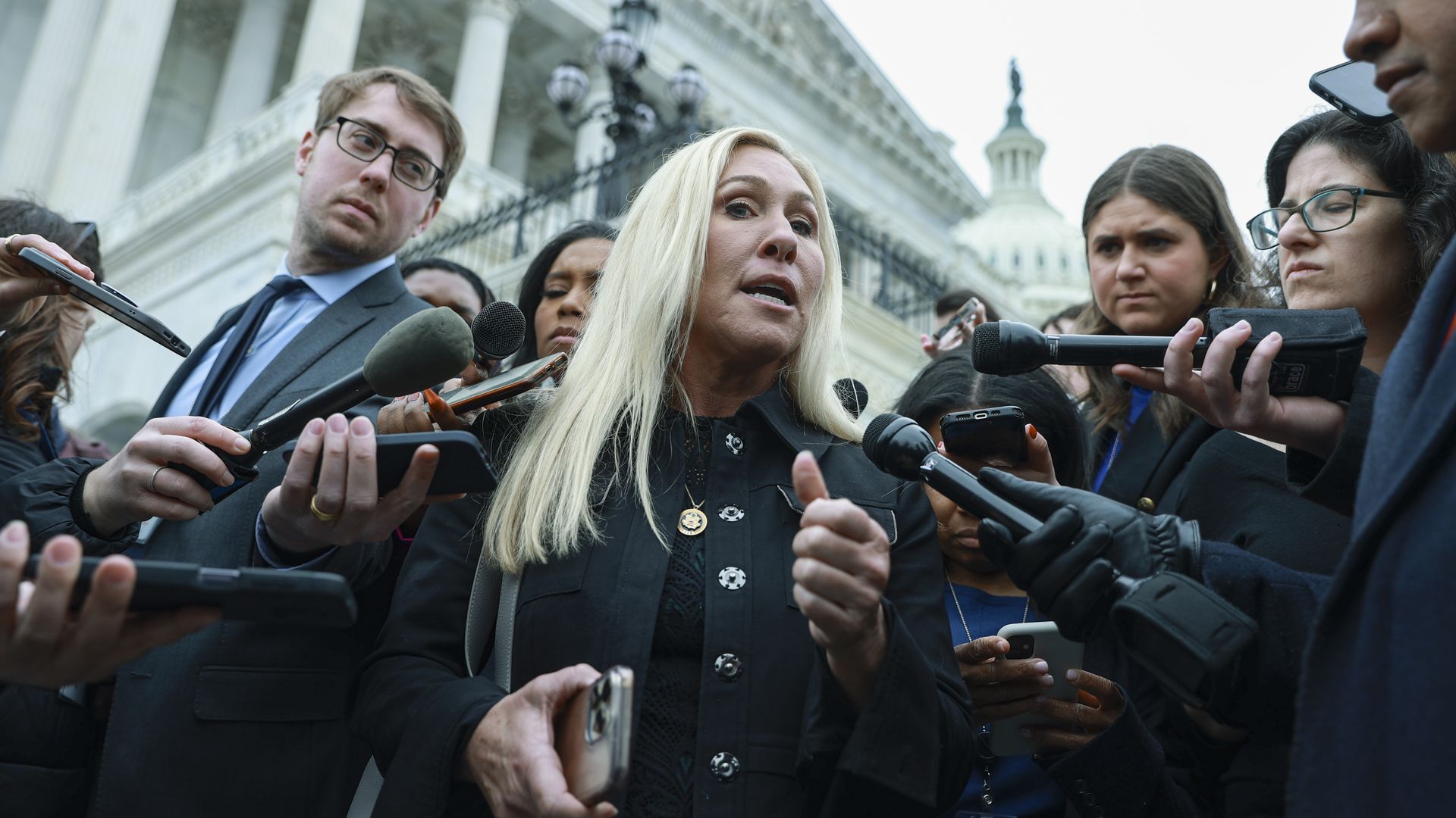 Rep. Marjorie Taylor Greene, wearing a black jacket and holding an iphone, speaks to reporters outside the Capitol.
