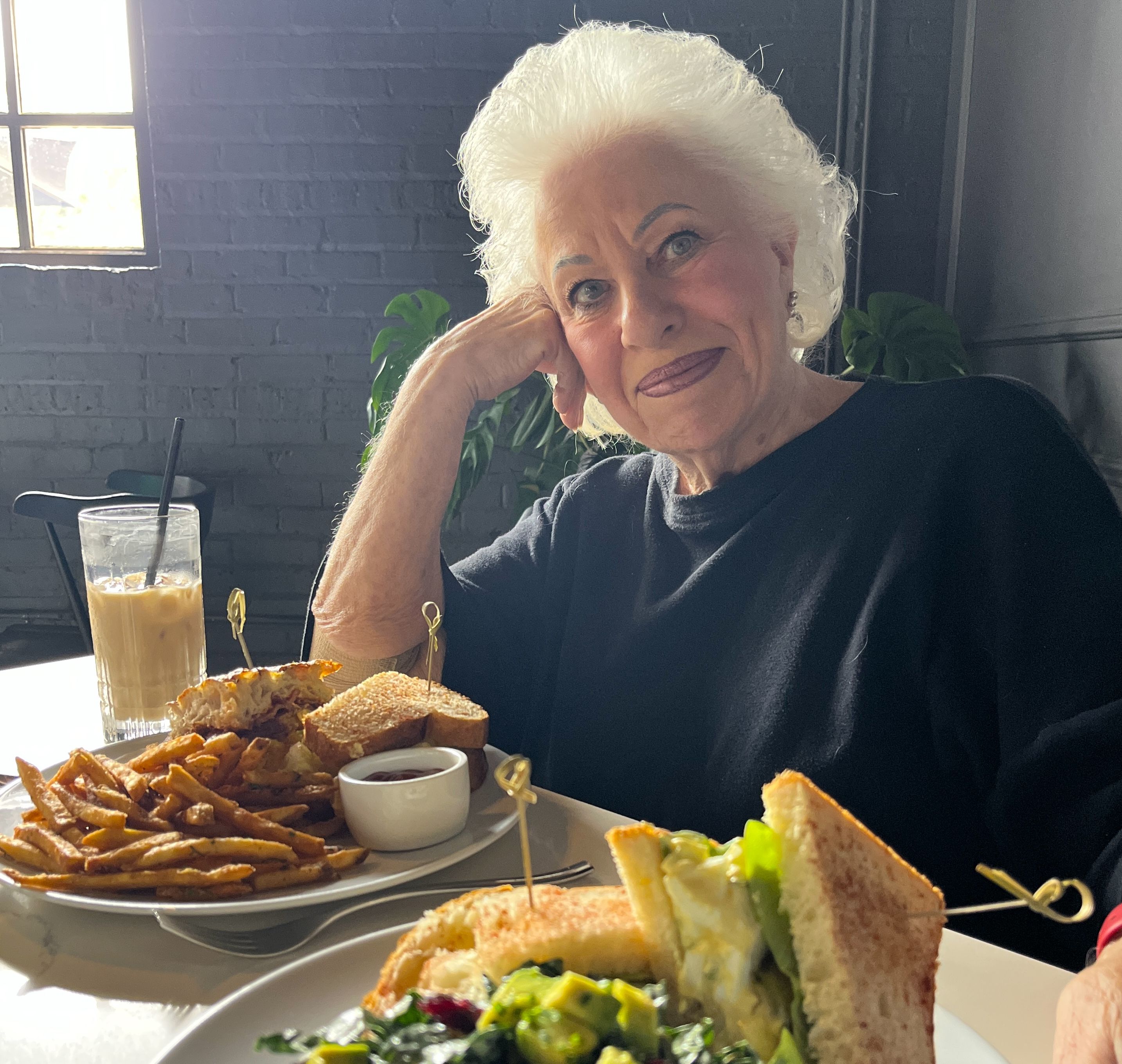 Elderly woman with white hair and black top sitting at a table with plates of sandwiches, fries, and a salad, and a glass of iced coffee in a cozy restaurant with gray brick walls.