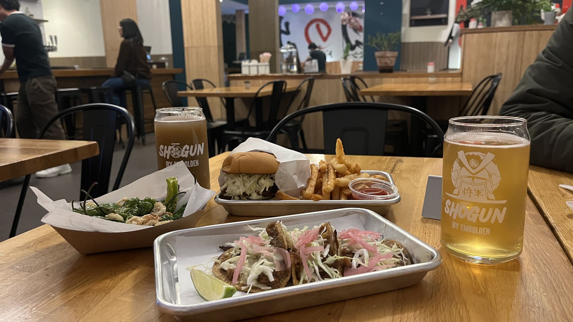 Table with food including three tacos with shredded cabbage and pink onions, a katsu sandwich with fries and ketchup, a tray of green shishito peppers, and two glasses of beer with "SHOGUN" branding in a casual restaurant.
