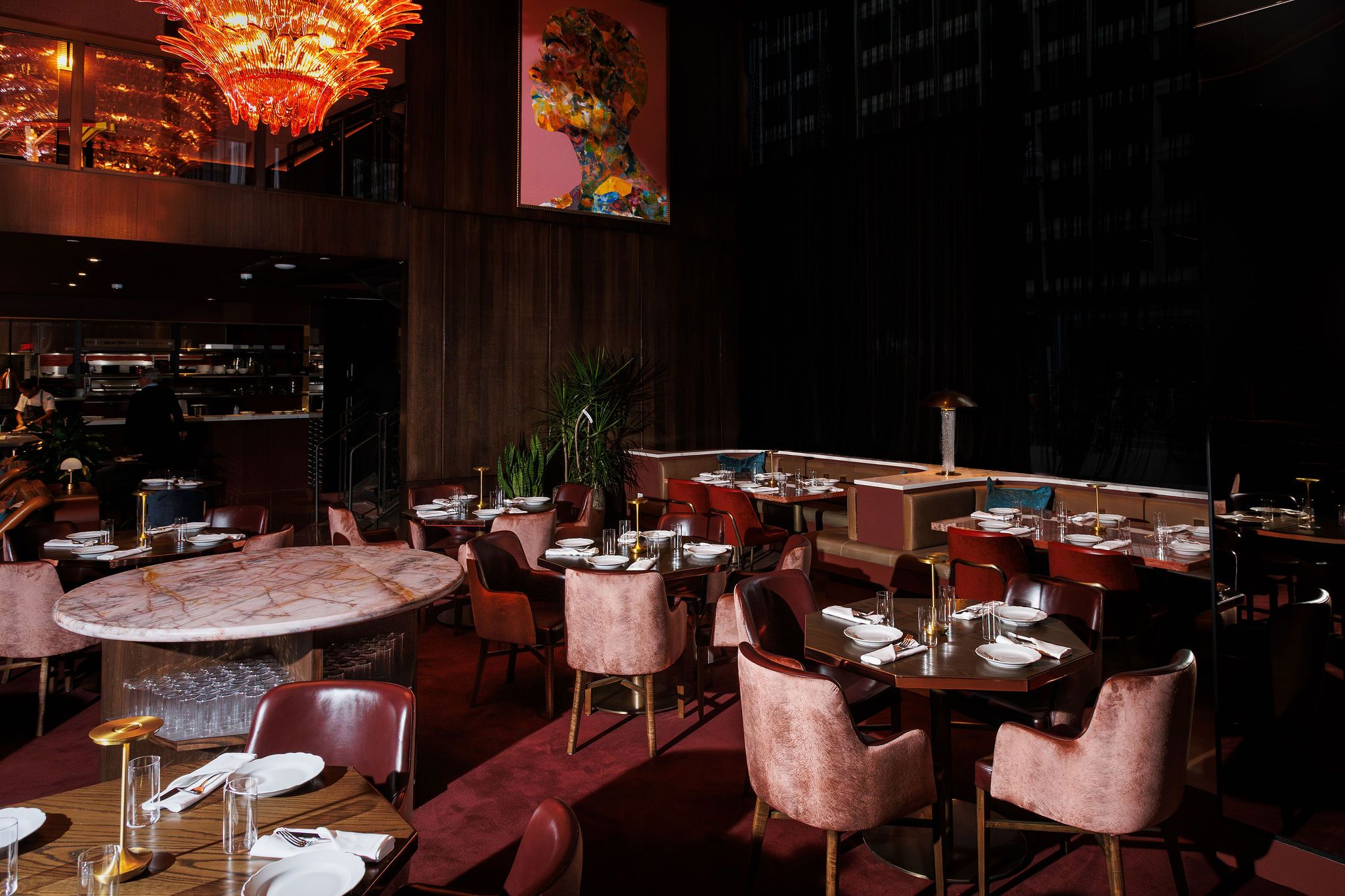 Dimly lit upscale restaurant with burgundy and pink velvet chairs, marble and wood tables set with white plates and glassware, warm amber chandelier, and colorful artwork on wood-paneled wall.