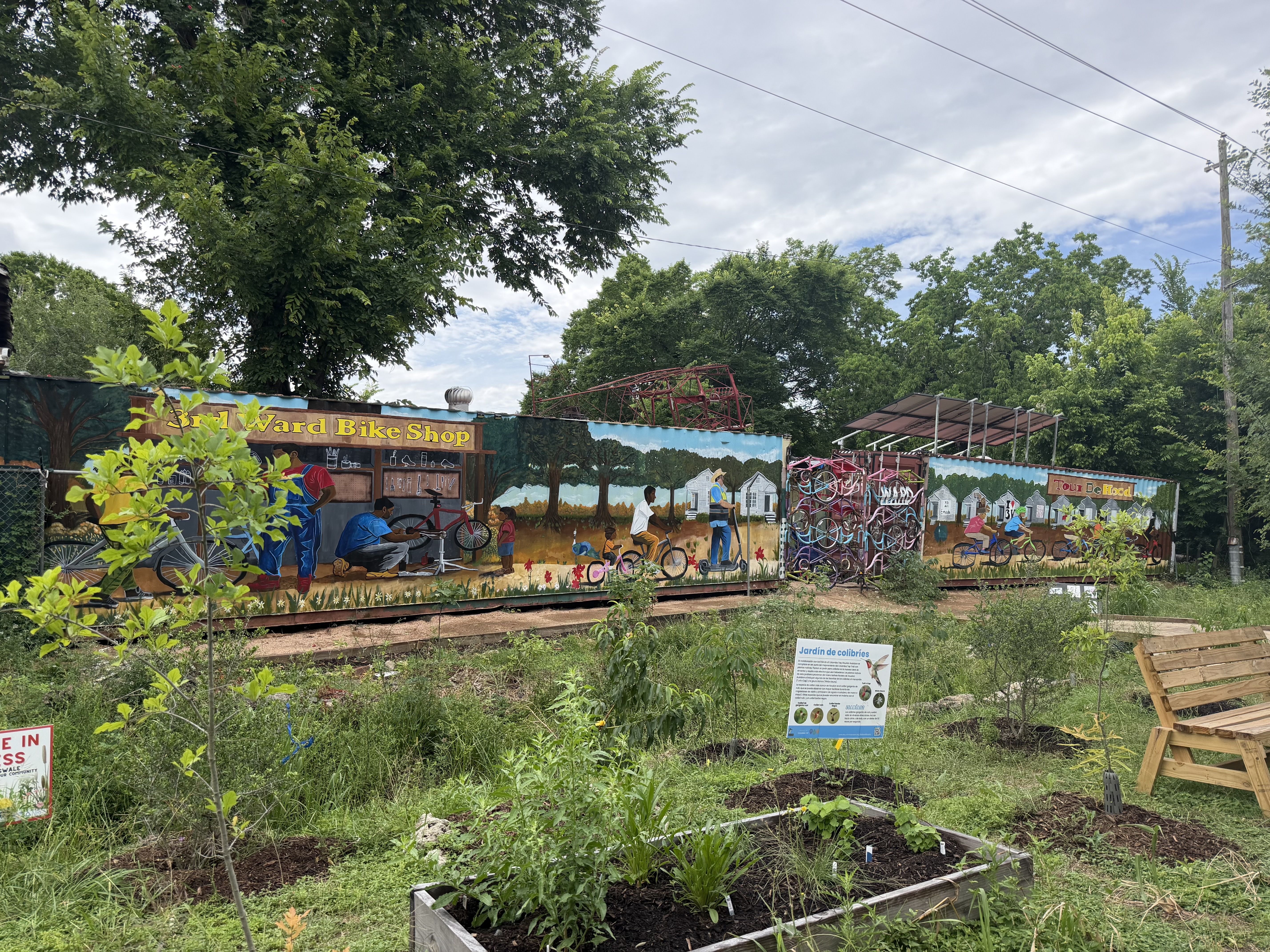 Outdoor mural along a fence shows people with bikes and a "Bike Shop" sign; tall green trees, power lines across a blue, cloudy sky, and raised garden beds with plants in the foreground.