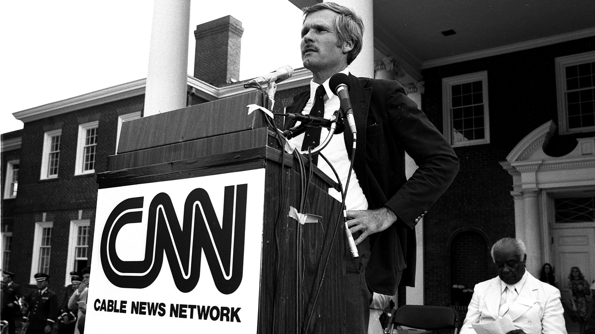 Ted Turner standing behind a podium with CNN Cable News Network attached to it. He's wearing a suit jacket and a tie and has his hand at his waist