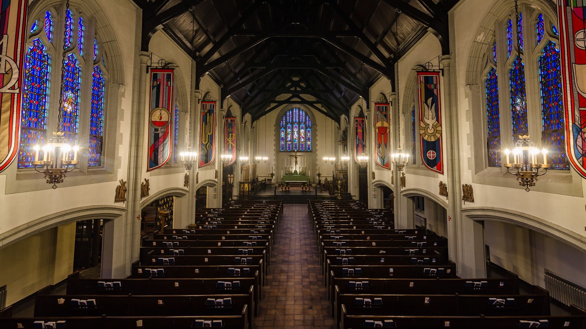 Interior of a church with wooden ceiling, stained glass windows in blue and red, hanging colorful banners, chandeliers, wooden pews, and an altar with candles and a crucifix.