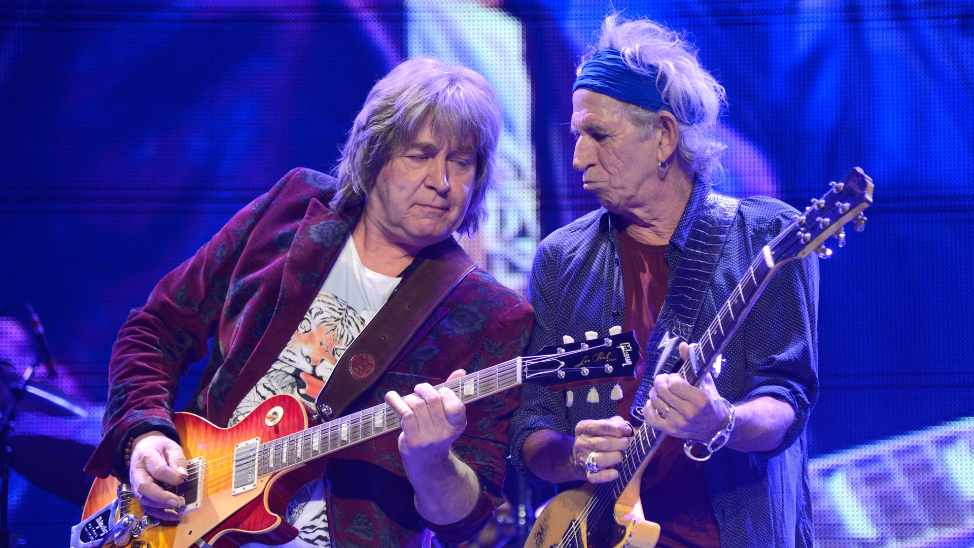 Two older male musicians play electric guitars on a blue-lit stage; one wears a red floral jacket and tiger tee, the other a dark shirt with a blue headband and multiple rings.