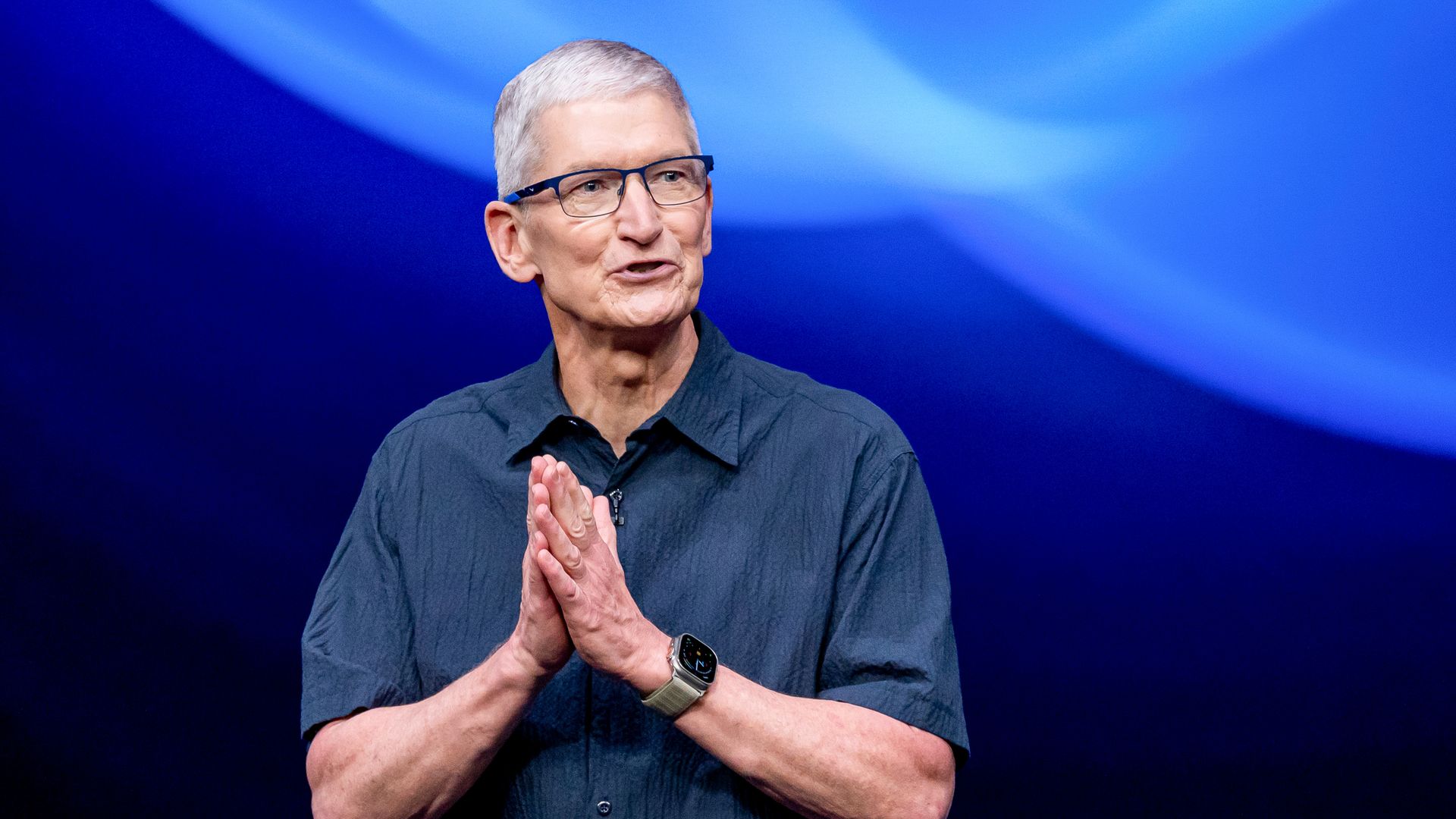 Middle-aged man with short gray hair and glasses speaking on stage wearing a dark blue short-sleeve shirt and a smartwatch, against a blue abstract background.