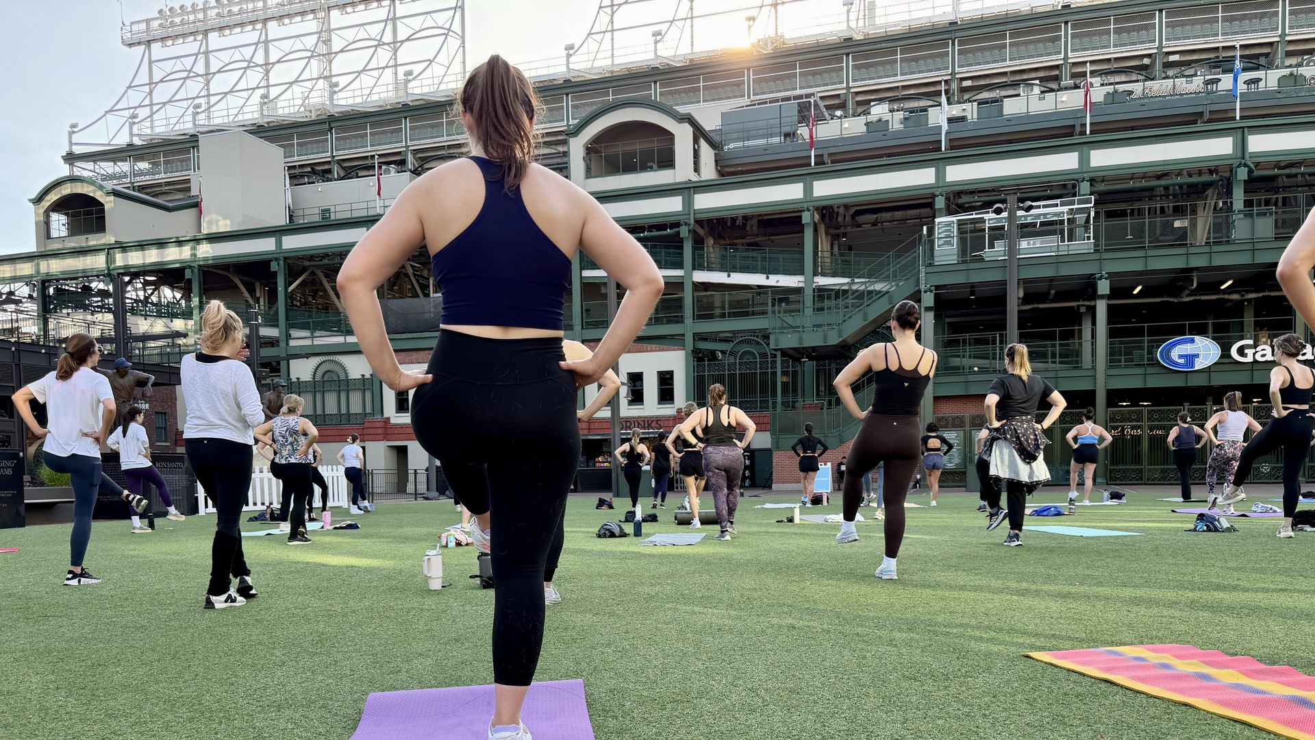 Rear view of a woman in a navy sports bra and black leggings leading a group workout on a purple mat, with a green stadium and other exercisers in the background.