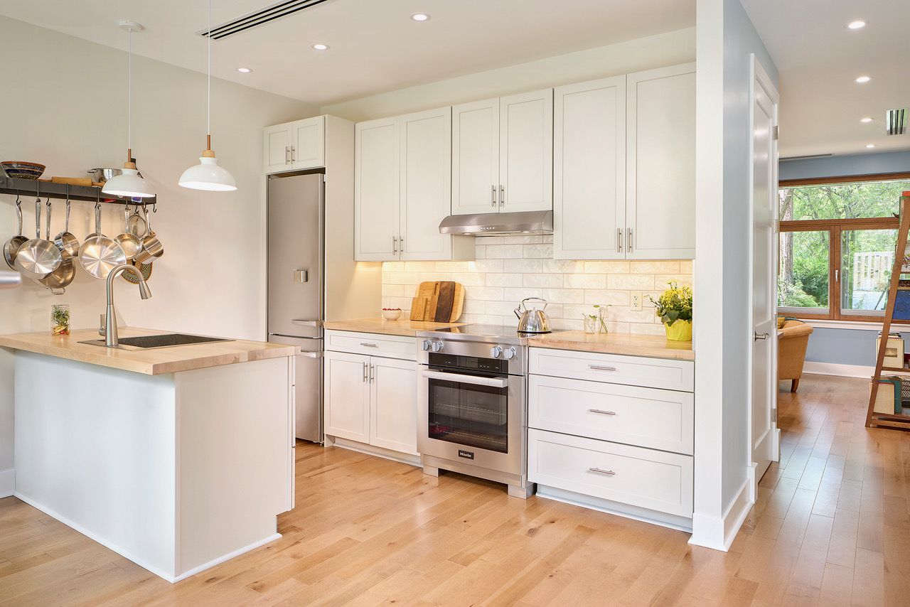 Bright modern kitchen with white cabinets, light wood countertops, stainless steel appliances, hanging pots, and pendant lights over a white island with a sink. Hardwood floor.