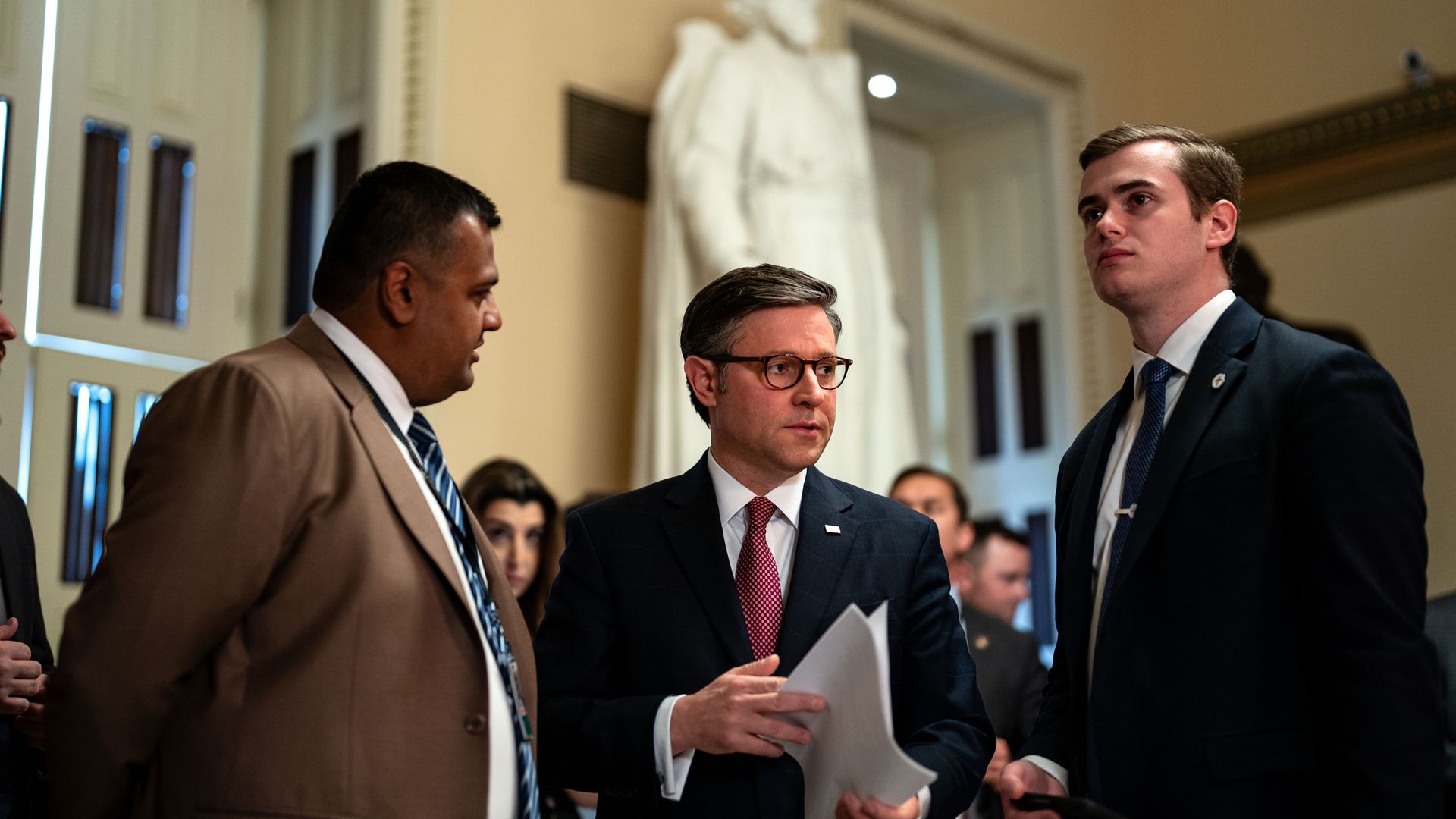 House Speaker Mike Johnson, wearing a gray suit, white shirt and red tie and flanked by aides in a Capitol hallway.