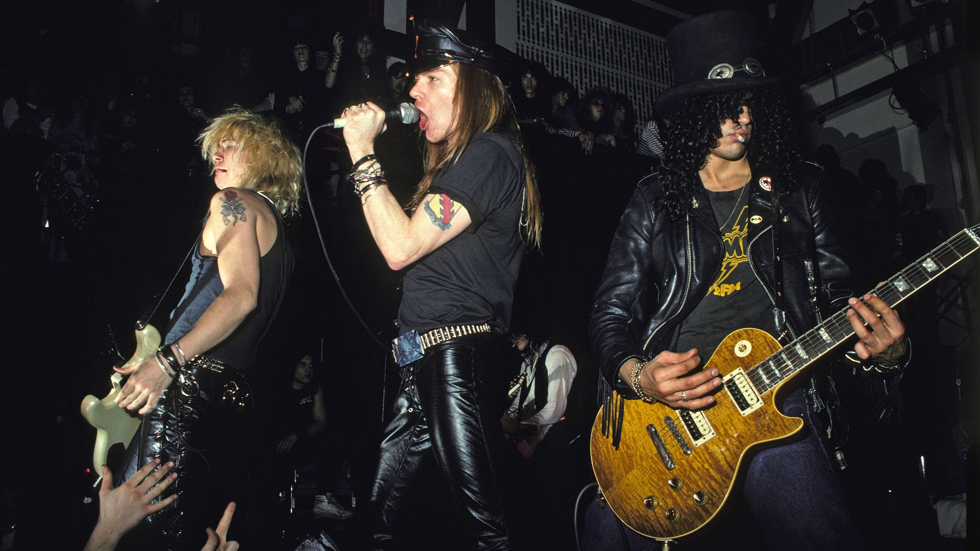 Three musicians from the rock band Guns N' Roses play on stage, a guitarist with tattoos and blonde hair on the left, a man wearing a black cap and leather pants singing into a microphone in the center, and a guitar player with a black top hat and long, curly hair on the right. 