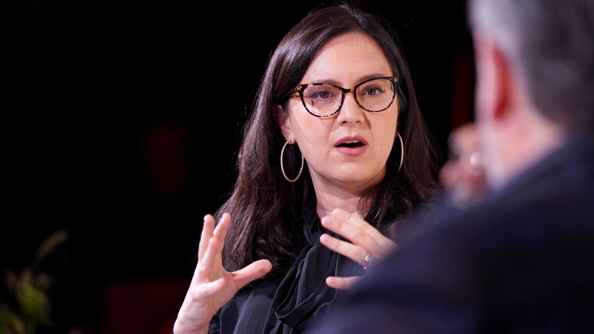 Bari Weiss, in glasses and with large hoop earrings, seated facing a speaker and gesturing with both hands open. 