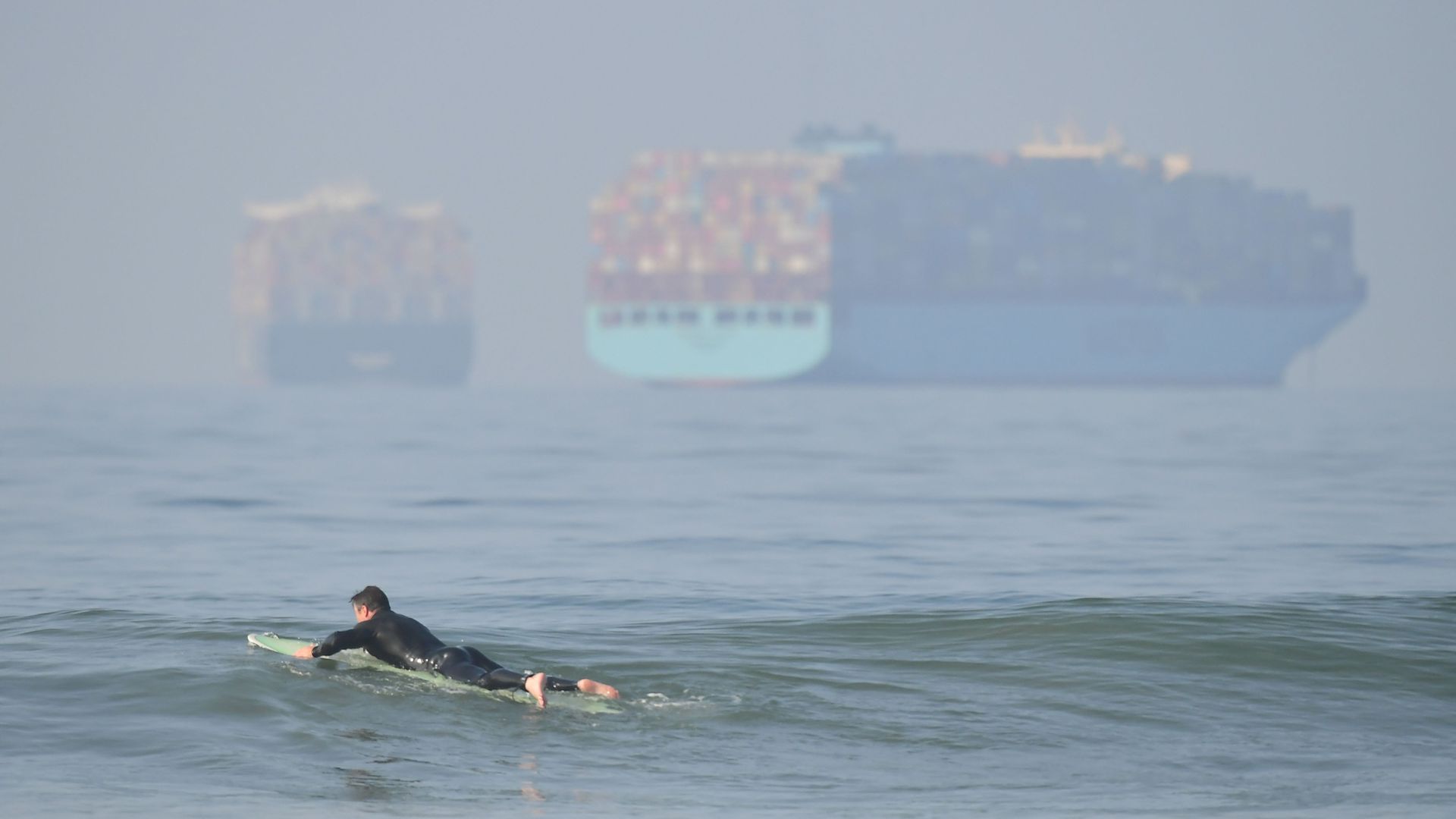 A surfer paddles in front of cargo ships