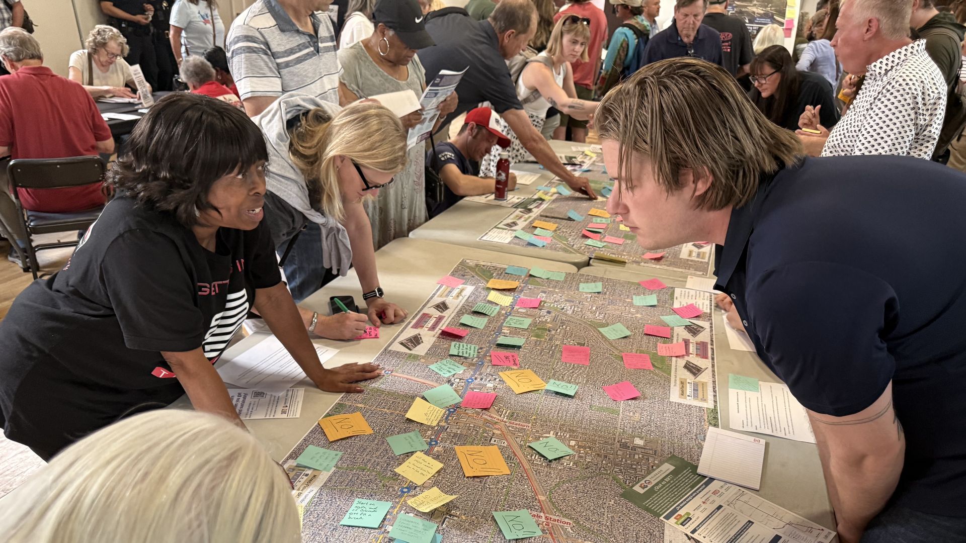 Crowded workshop around a long table with a large city map covered in colorful Post-it notes. Foreground shows a woman with short black hair and a man with long blond hair studying the board.