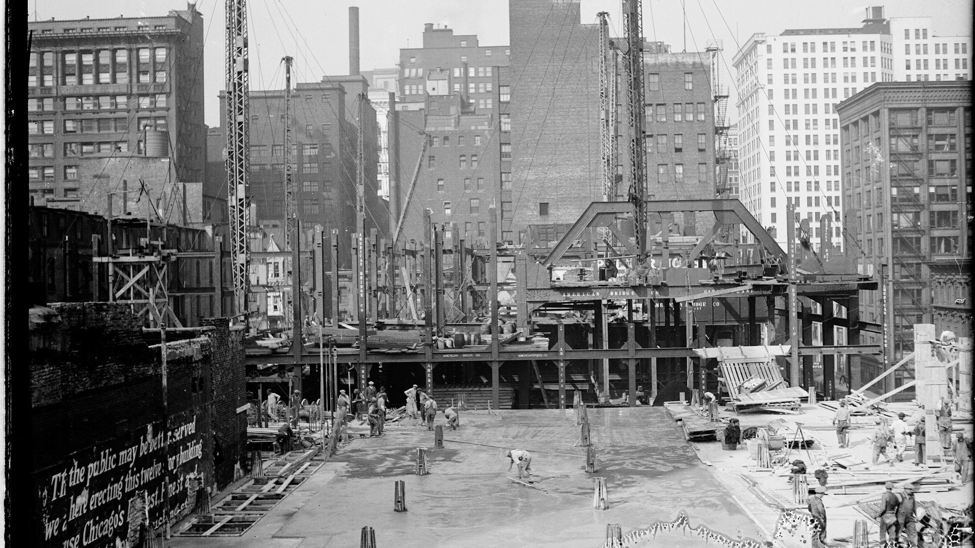 Black-and-white city construction scene with workers on the ground among scaffolding and tall cranes, brick buildings rising in the background.