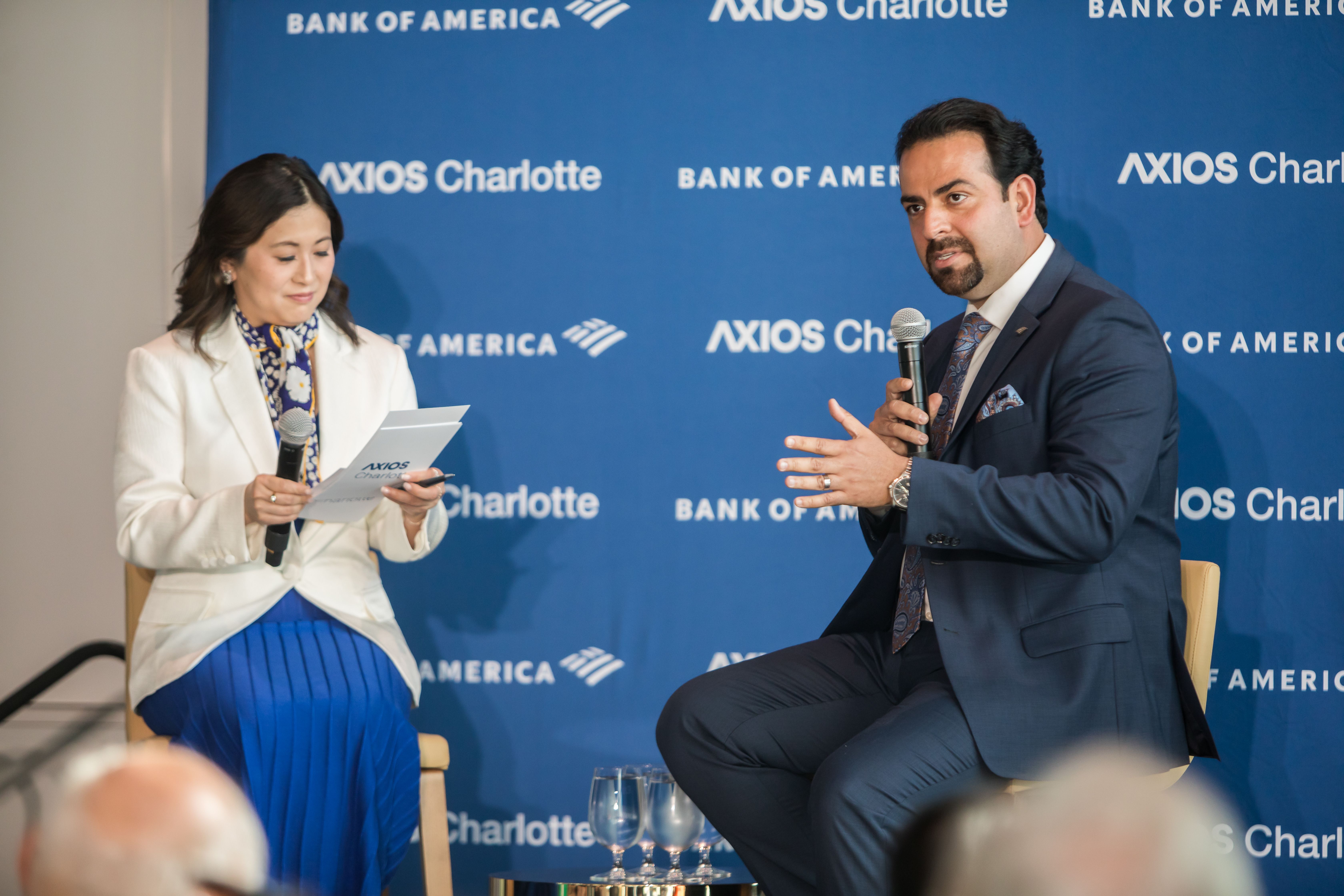 Two speakers on a stage in front of a blue backdrop with Axios Charlotte and Bank of America logos; a woman in a white blazer reads notes, while a man in a dark suit speaks into a microphone.