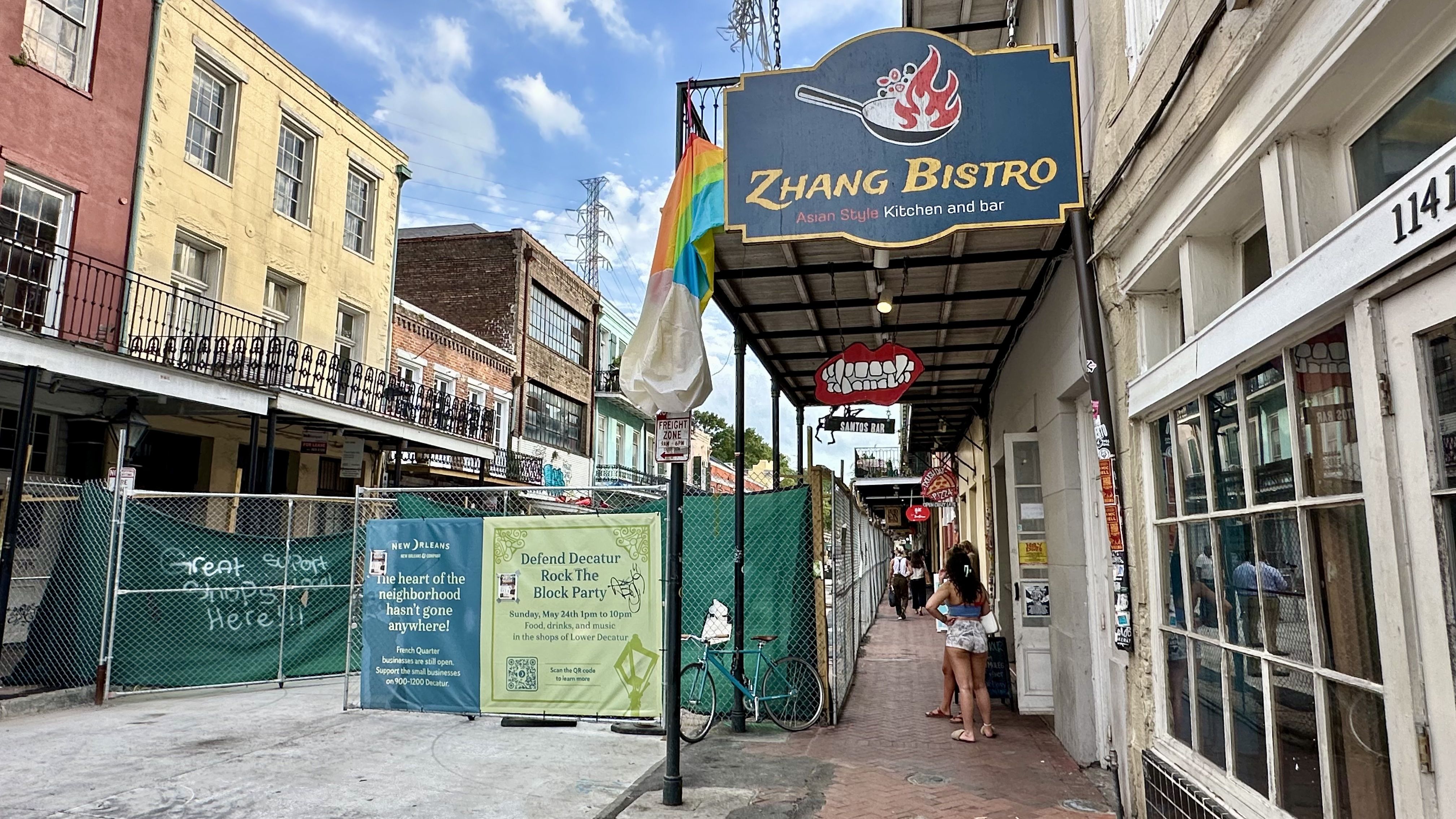 Sunny urban alley with a blue Zhang Bistro sign; rainbow flag nearby. A person in shorts walks along a covered sidewalk, flanked by colorful buildings and a chain-link fence.