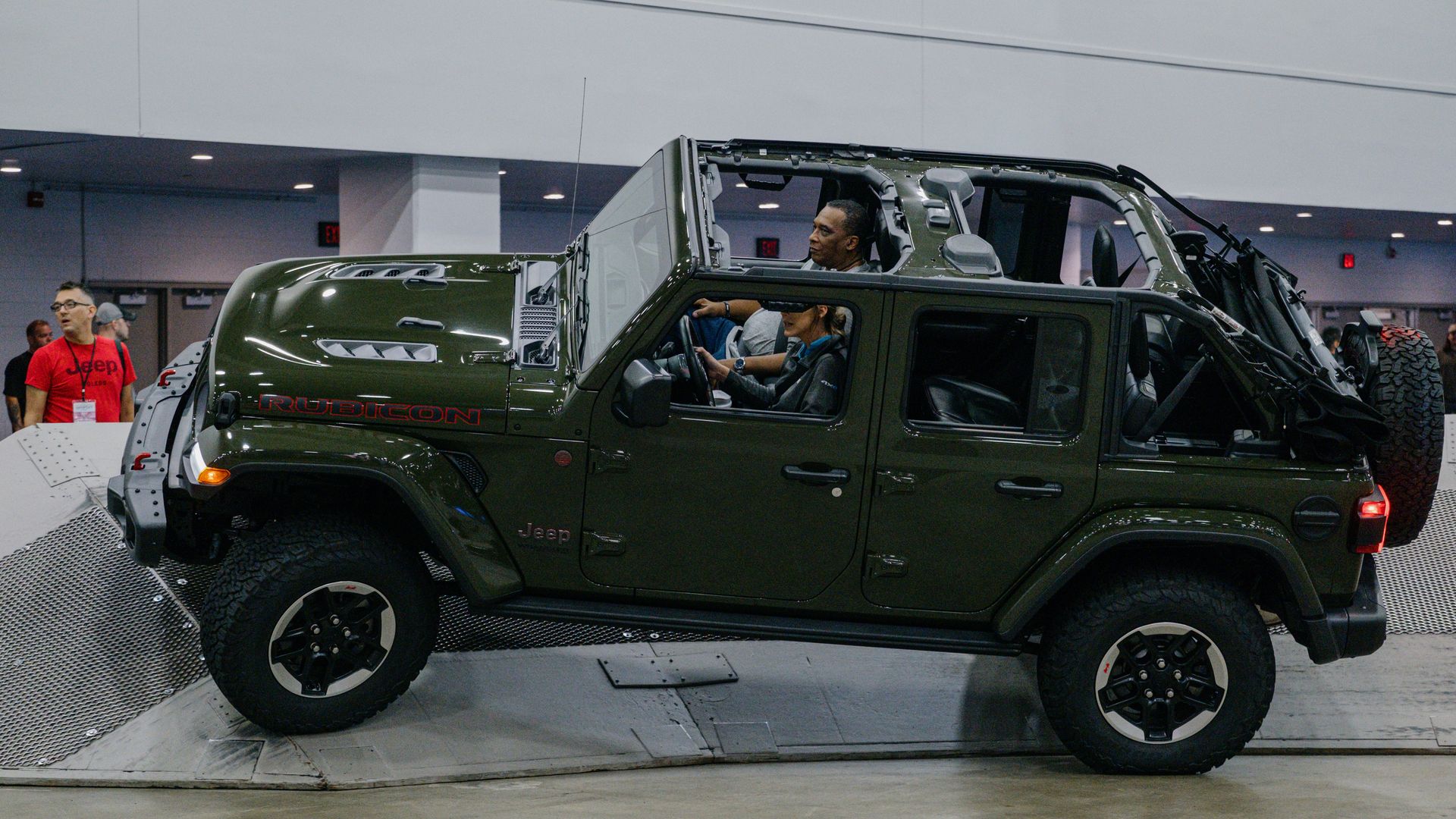 Dark green Jeep Wrangler Rubicon with roof removed on a showroom display; two people sit inside while attendees observe at an indoor expo.