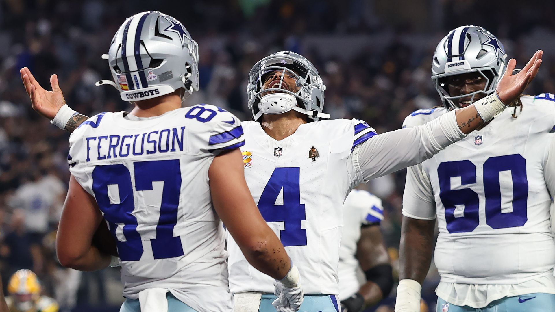 Dak Prescott, surrounded by his teammates, raises both hands in the air while smiling and looking up