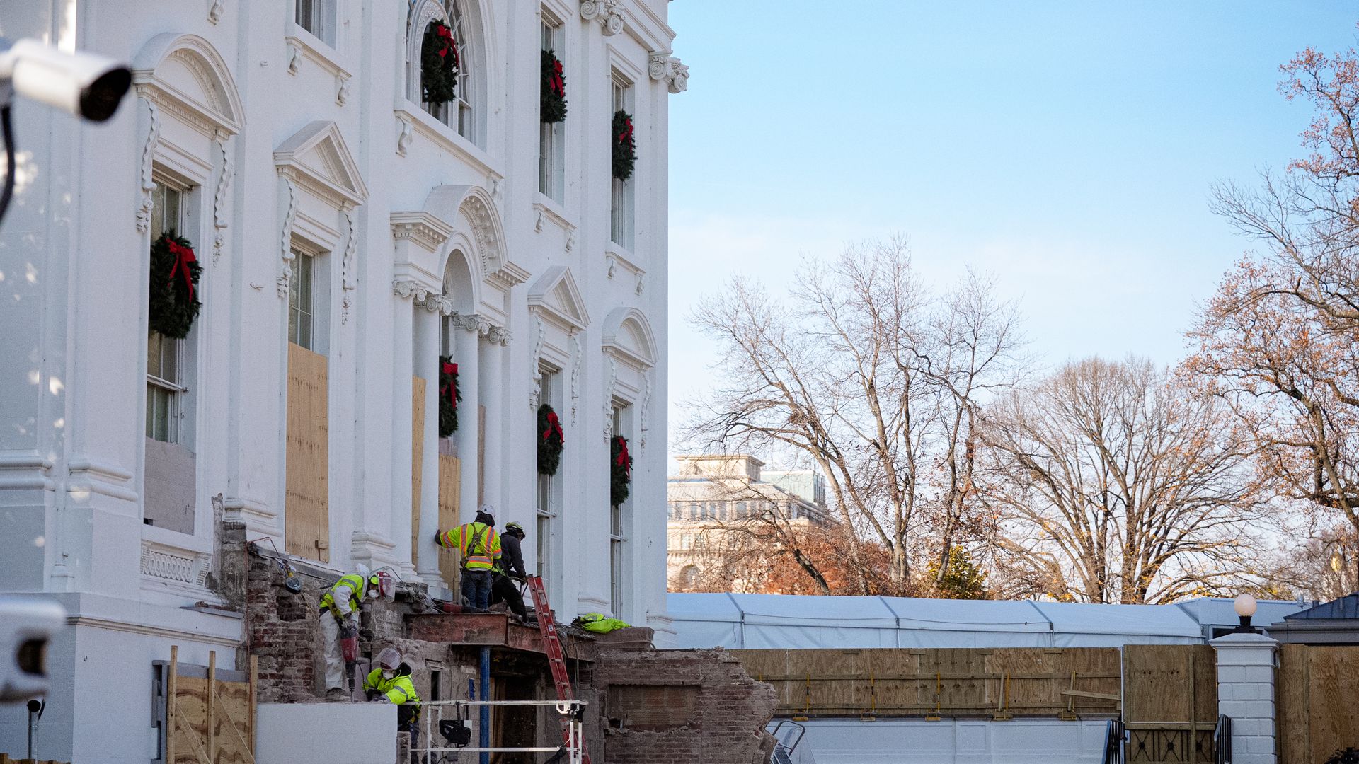 Workers in yellow safety vests repair a white building decorated with Christmas wreaths with red bows, some windows boarded up, under a clear blue sky with leafless trees nearby.