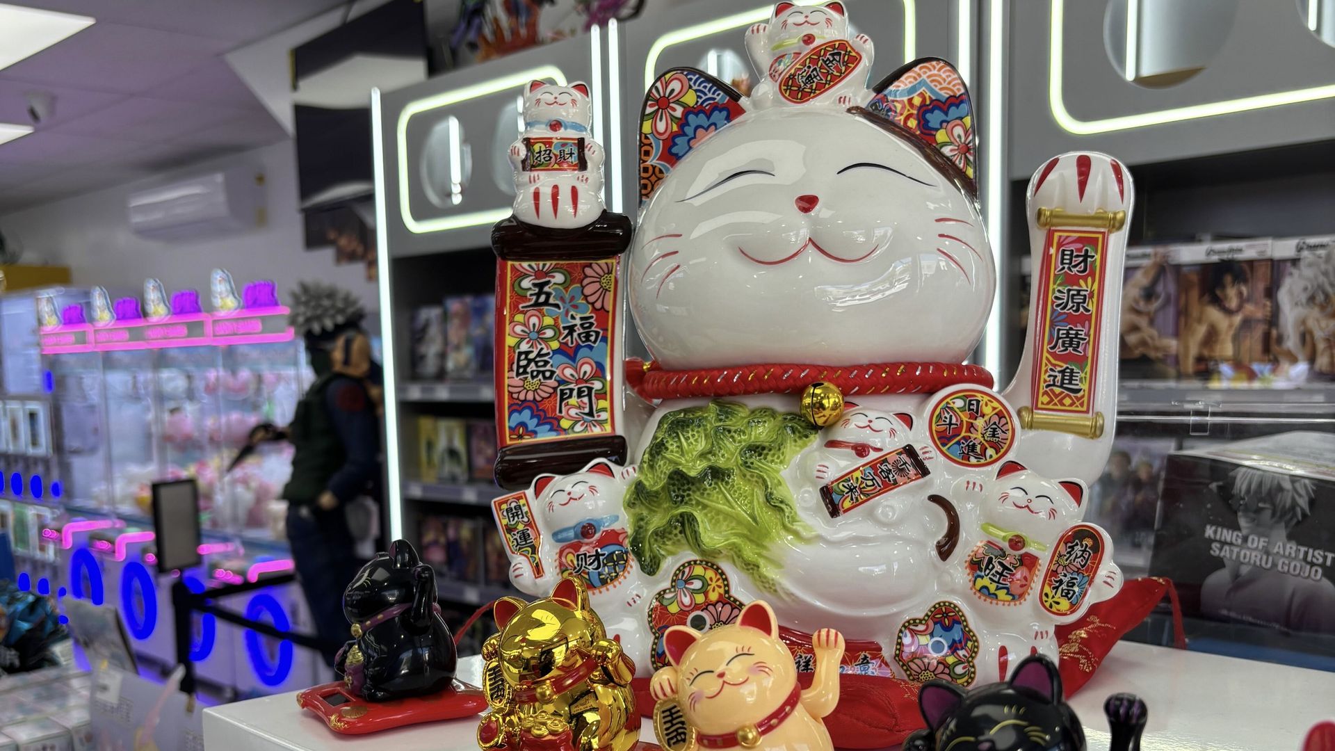 Large white ceramic lucky cat with colorful floral patterns and smaller cats on it, surrounded by smaller black, gold, and orange lucky cat figurines, displayed in a brightly lit store.