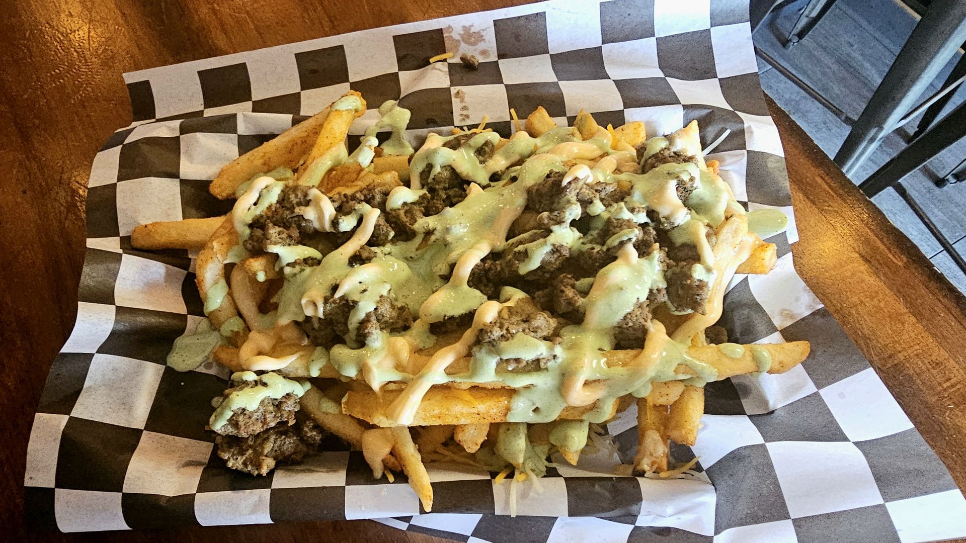 Pile of thick fries loaded with seasoned ground beef, melted cheese, and green and creamy sauces, served on black-and-white checkered parchment over a wooden table.