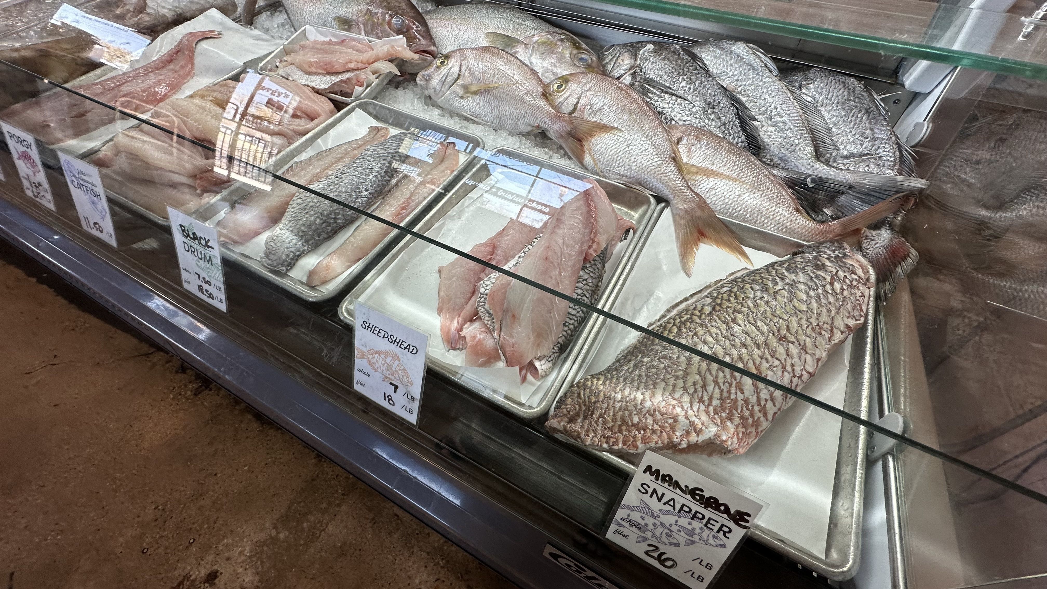 A zoomed-in look at a glass seafood display case. Cut fillets are advertised as mangrove snapper, sheepshead, black drum and catfish.