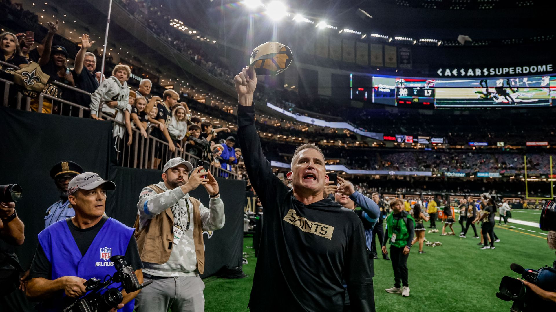 Darren Rizzi raises a Saints ball cap toward fans in the Caesars Superdome stands as he exits the field.