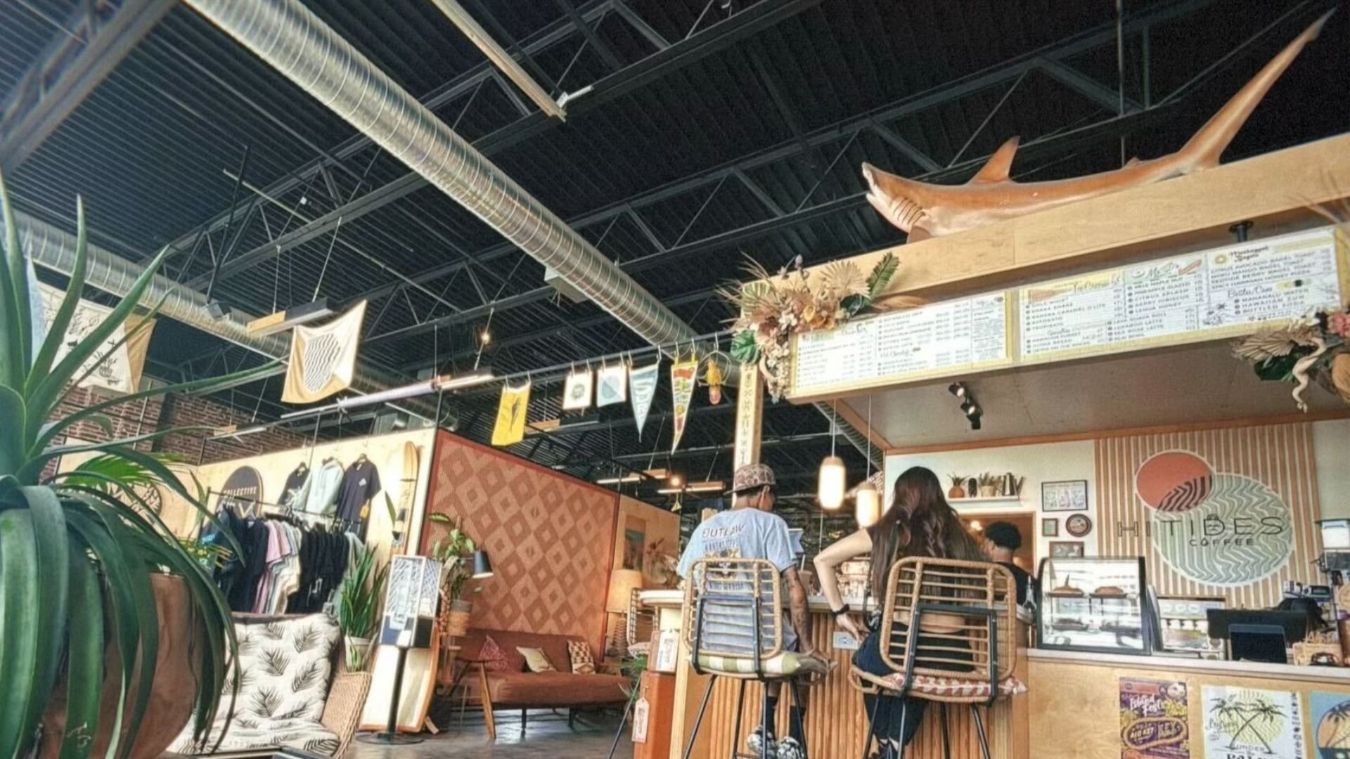 Interior of a cozy coffee shop with wooden decor, a large menu board, hanging flags, a mannequin shark above the counter, and two people seated on stools facing the bar.